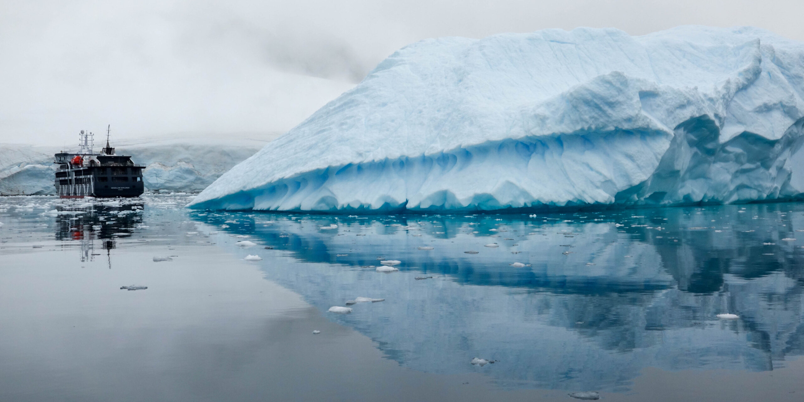 Expedition ship dwarfed by a massive, blue iceberg in the calm, gray waters of Antarctica.