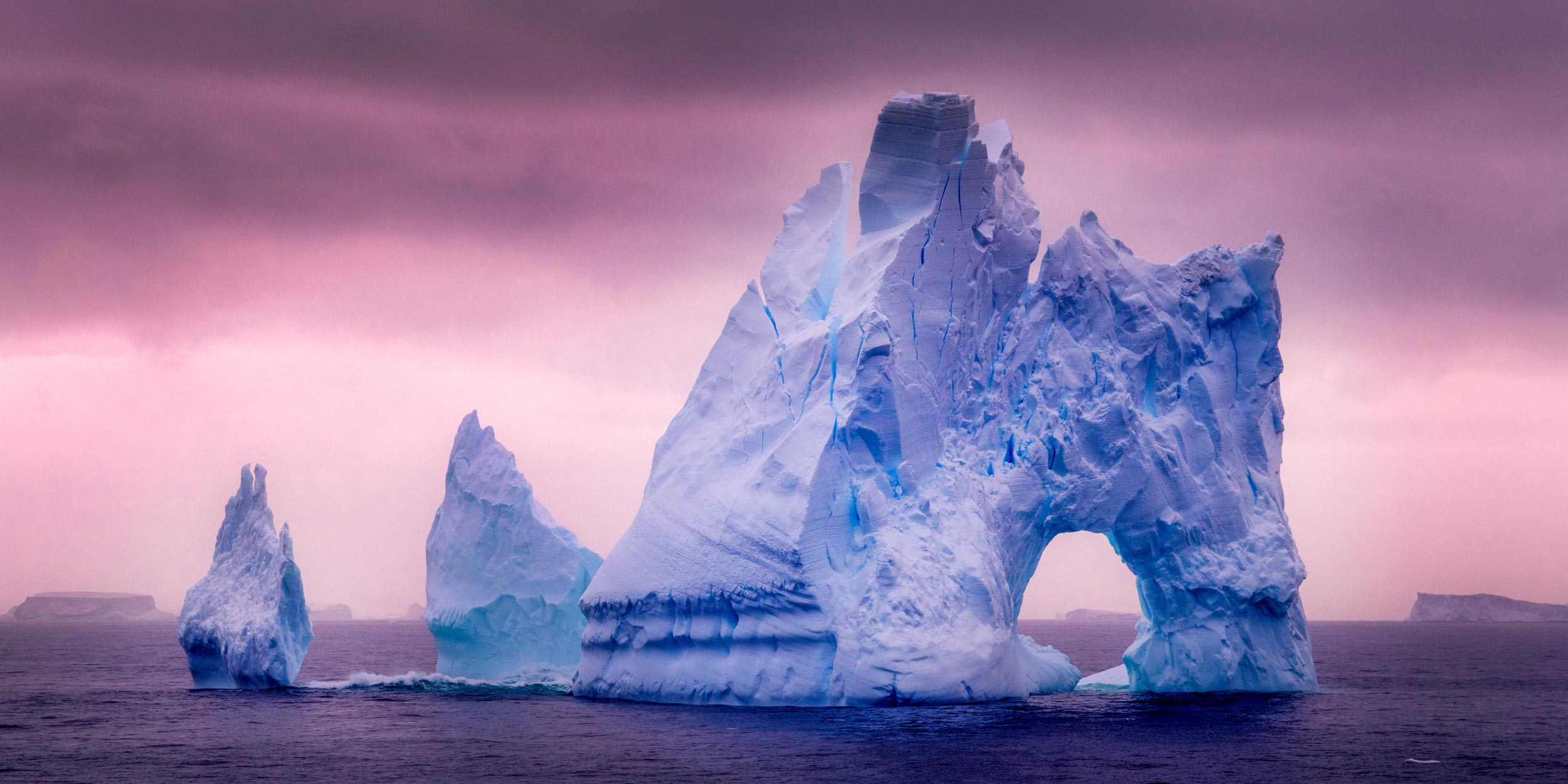 Sculptural blue iceberg with an arch and a smaller berg in dark water under a pink and purple sky.