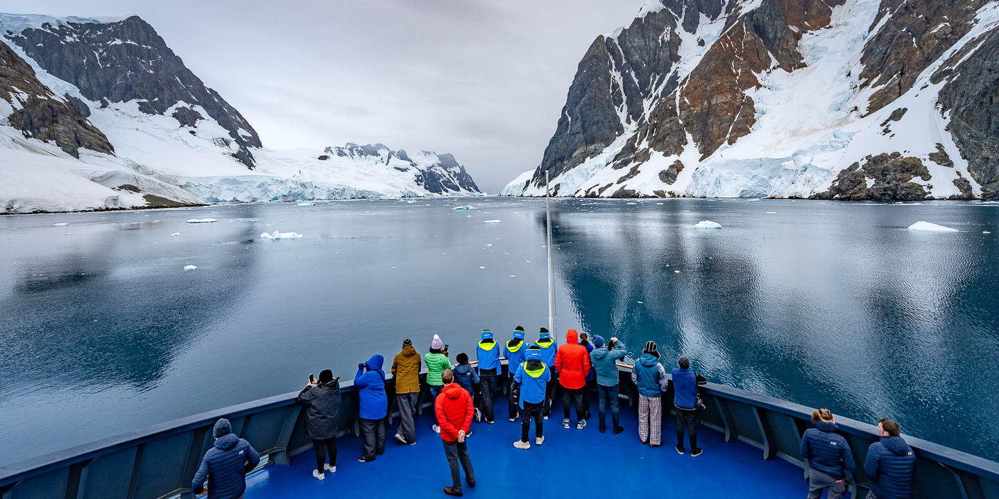 Expedition passengers standing on the bow of a ship viewing a glacial bay surrounded by mountains.