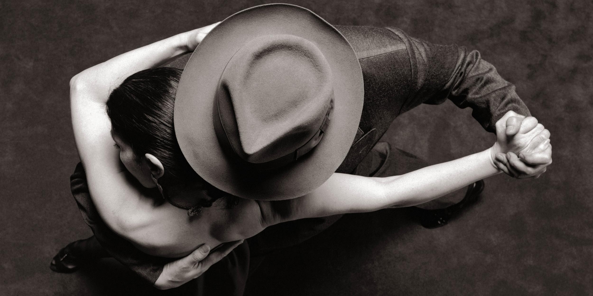 Black and white, overhead view of a couple dancing tango; the man wears a hat covering the woman's face.