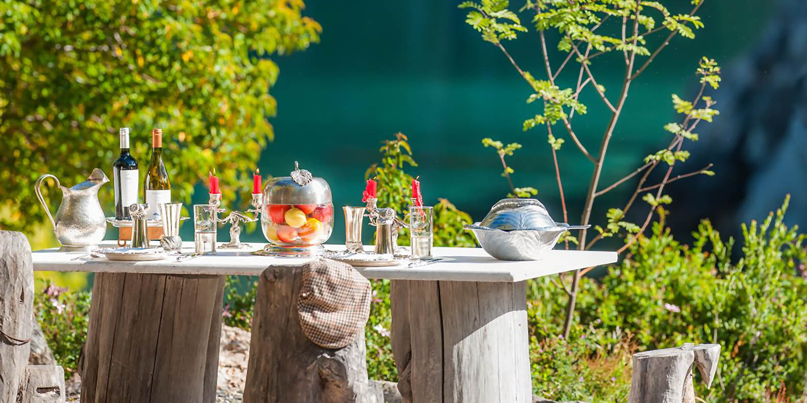 Outdoor dining table with silver service, candles, wine, and a fruit bowl by a bright blue lake.