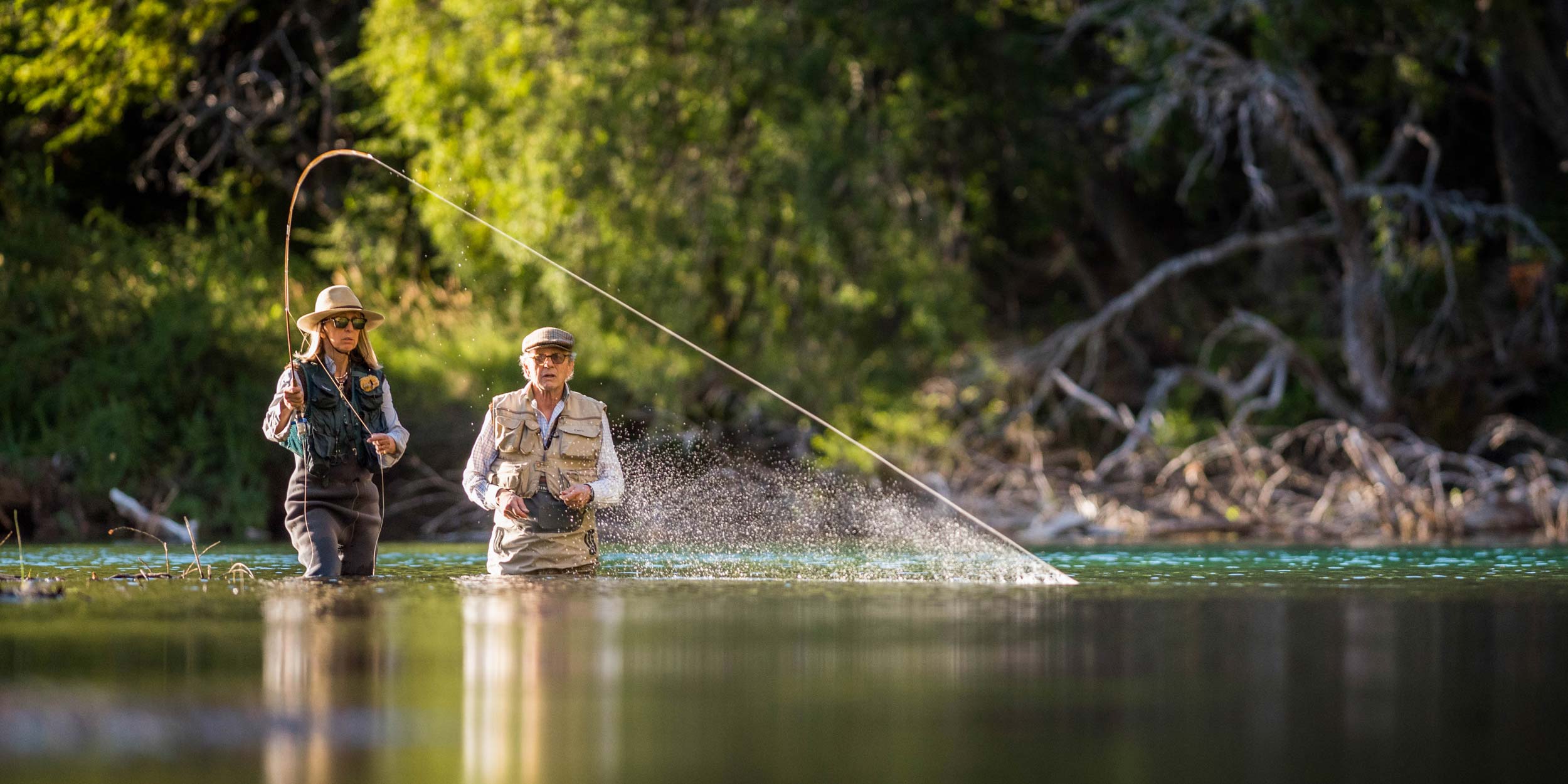 A man and woman fly fishing, standing waist-deep in a river, with green forest lining the banks.