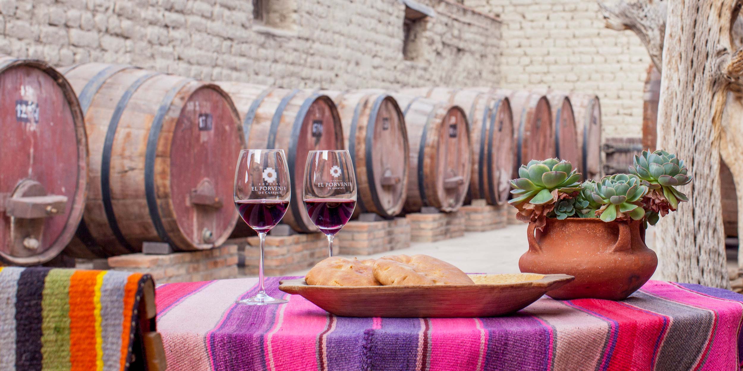 Two glasses of red wine and bread on a colorful table in front of wooden wine barrels at a Mendoza winery.