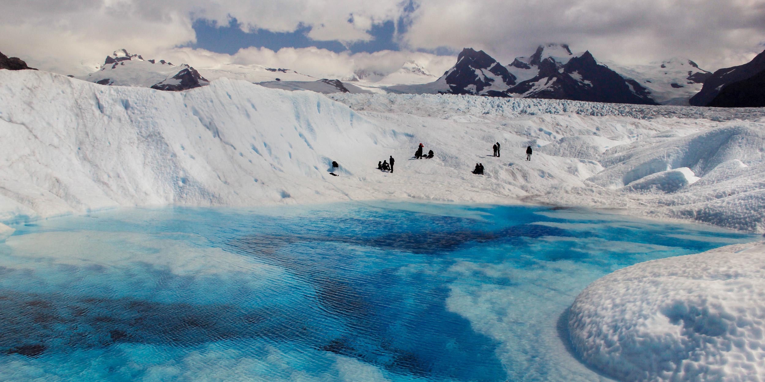 Tourists stand on the vast, white Perito Moreno Glacier next to a bright turquoise meltwater pond.