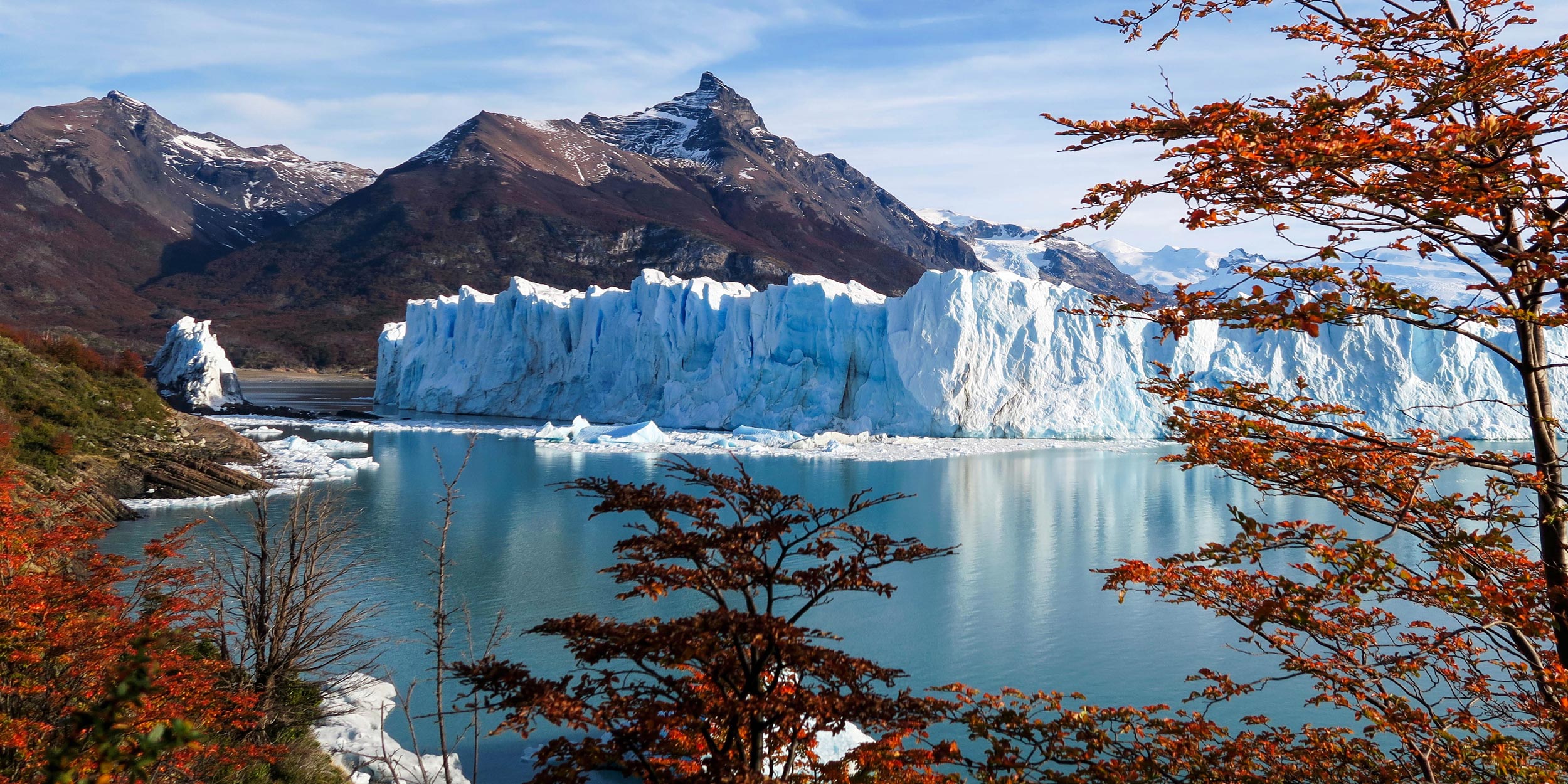 The massive, bright blue wall of the Perito Moreno Glacier meets a lake, framed by mountains and fall trees.