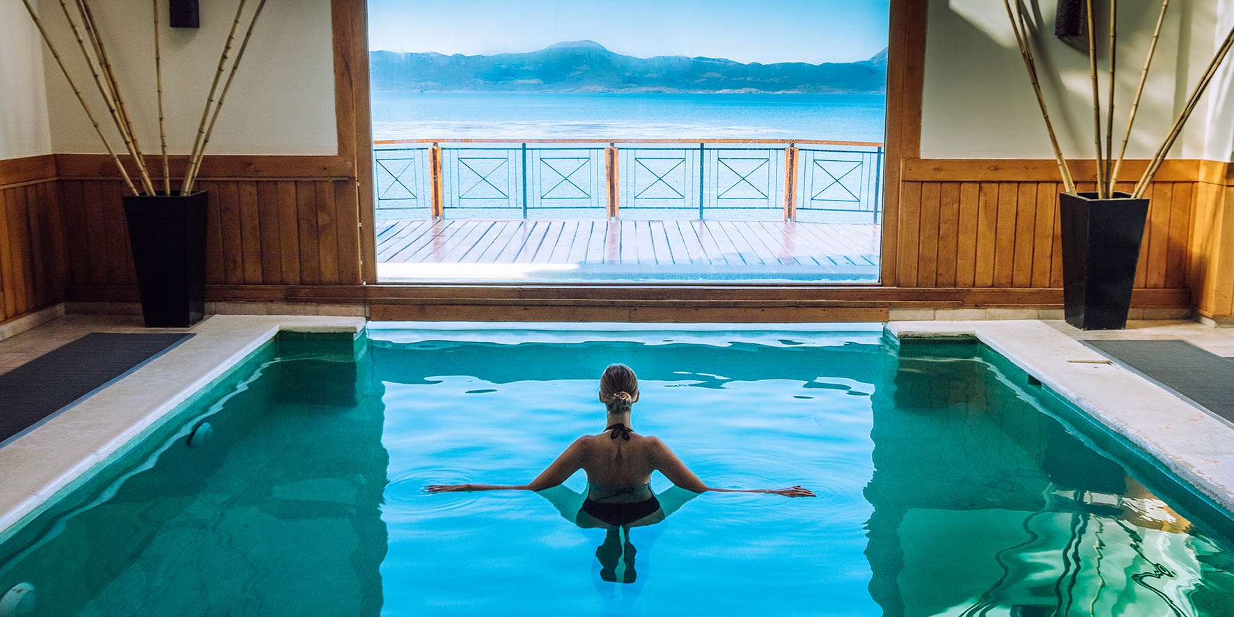 A woman relaxes in a bright indoor pool with an open deck providing a stunning view of a Patagonian lake.