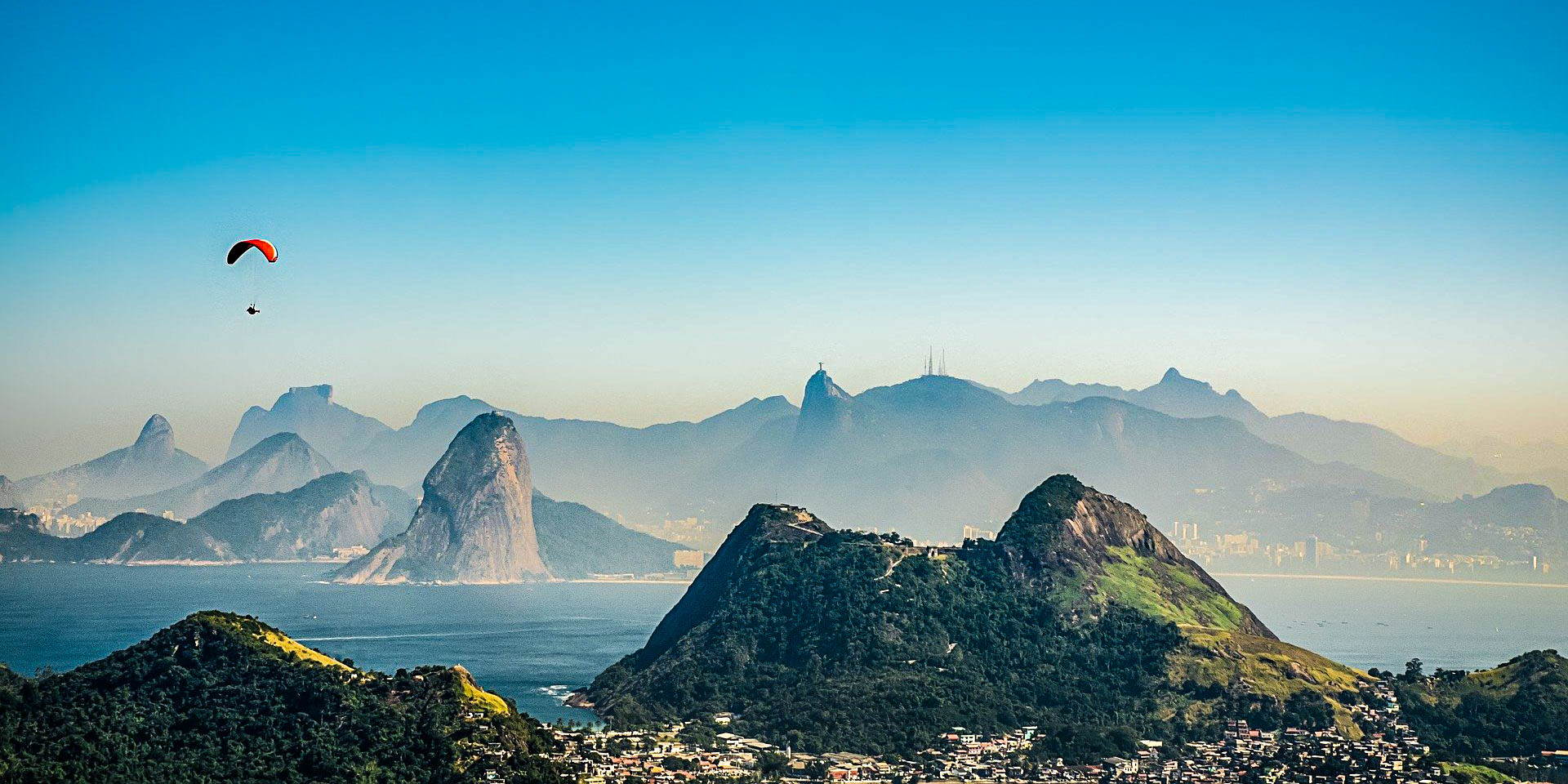 Paraglider soaring above Rio de Janeiro's mountainous coastline, with Sugarloaf and Christ the Redeemer visible.
