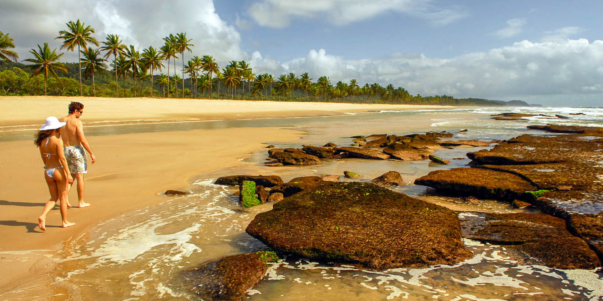 Couple walking along a secluded Brazilian beach with palm trees and large, dark, moss-covered rocks.