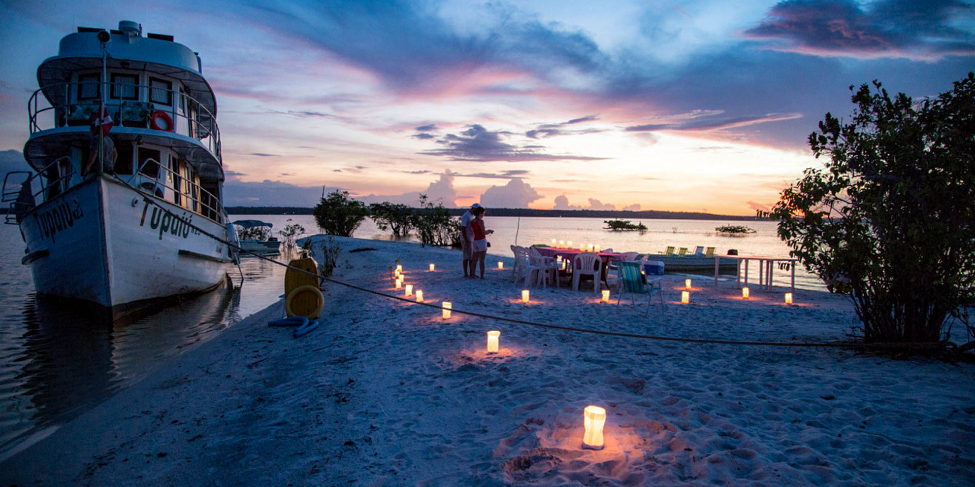 Candlelit private dinner on a sandy bank of the Tapajós River next to a moored boat at sunset.
