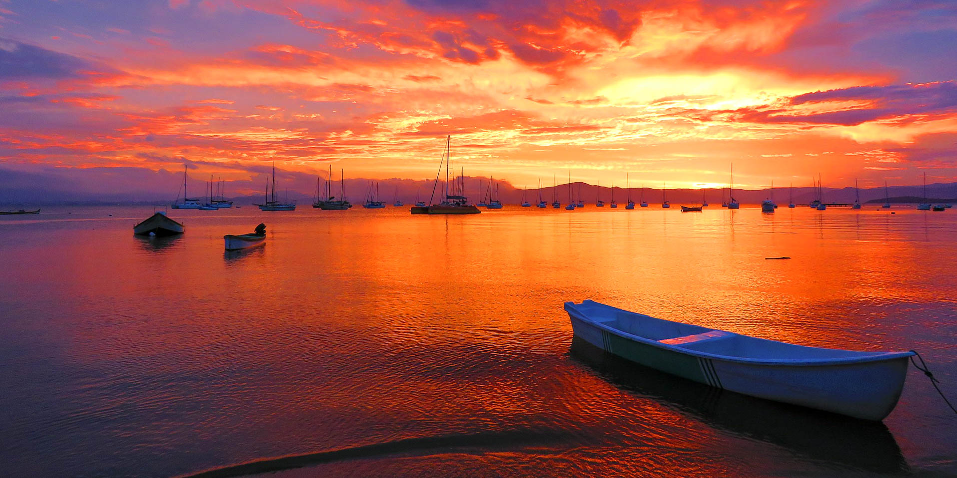 Vibrant orange and yellow sunset over the water in Buzios, Brazil, with boats anchored in the distance.