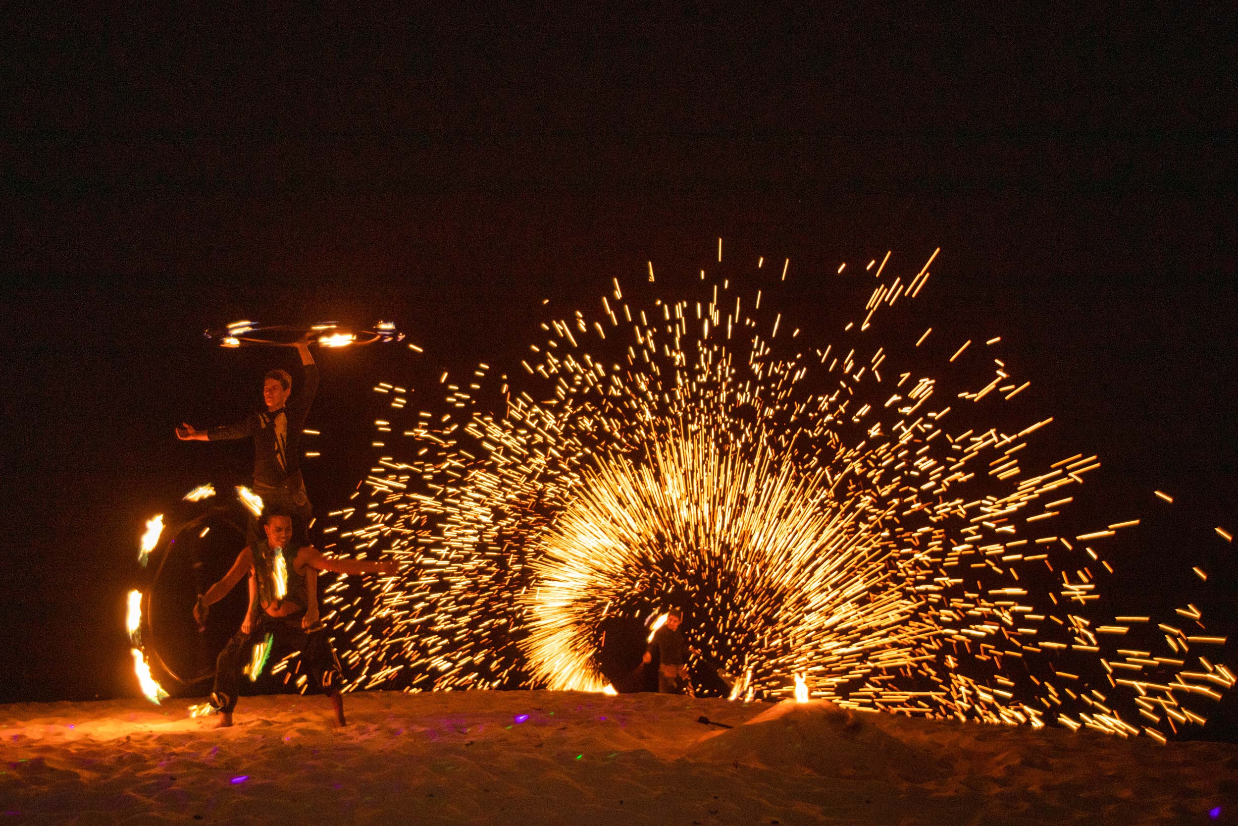 Fire dancers perform on a beach with a fireworks display in the background.