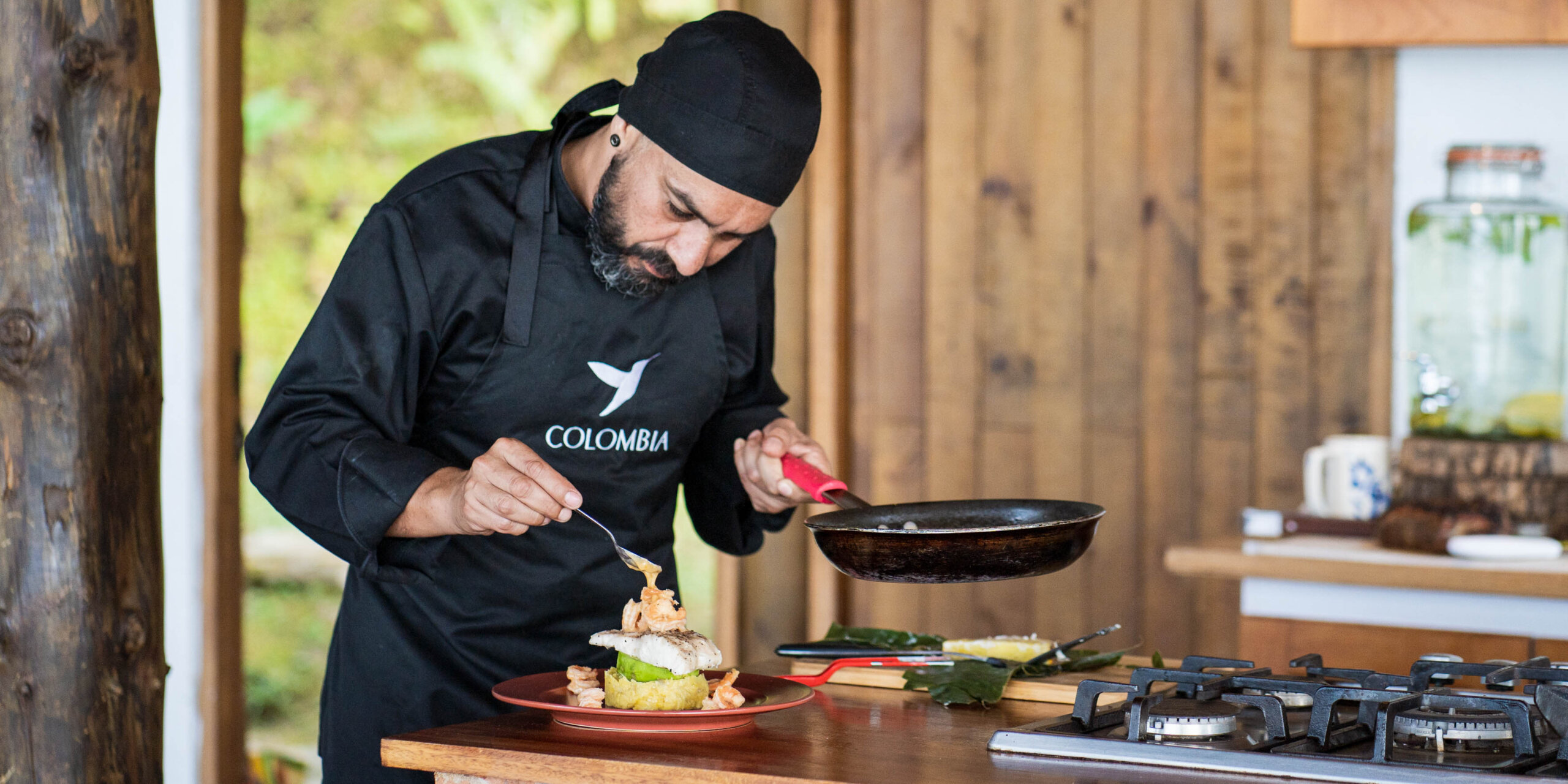 Chef in a black apron preparing a dish with shrimp and fish on a wooden counter.