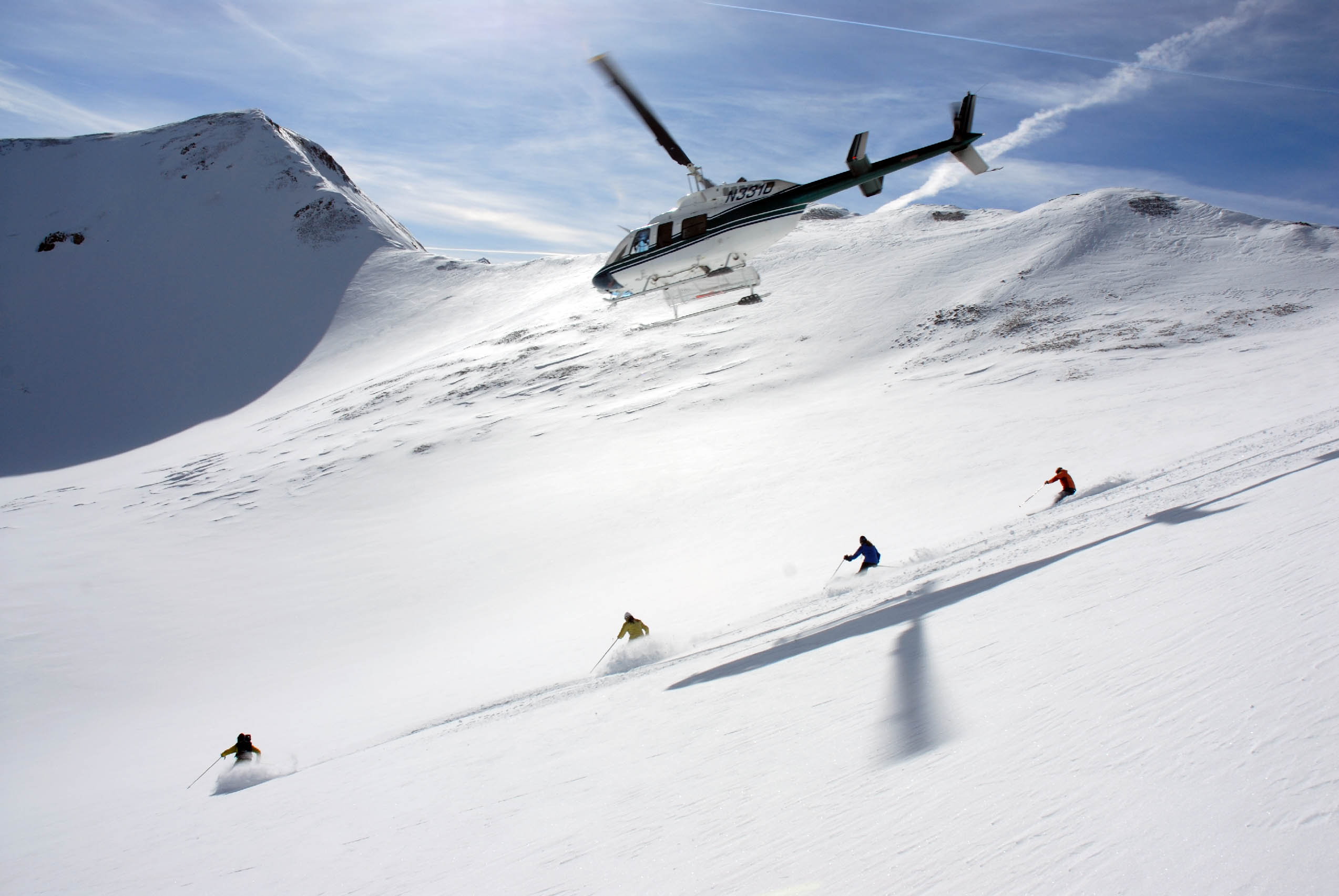 Skiers race down a mountain with a helicopter overhead in Colorado.