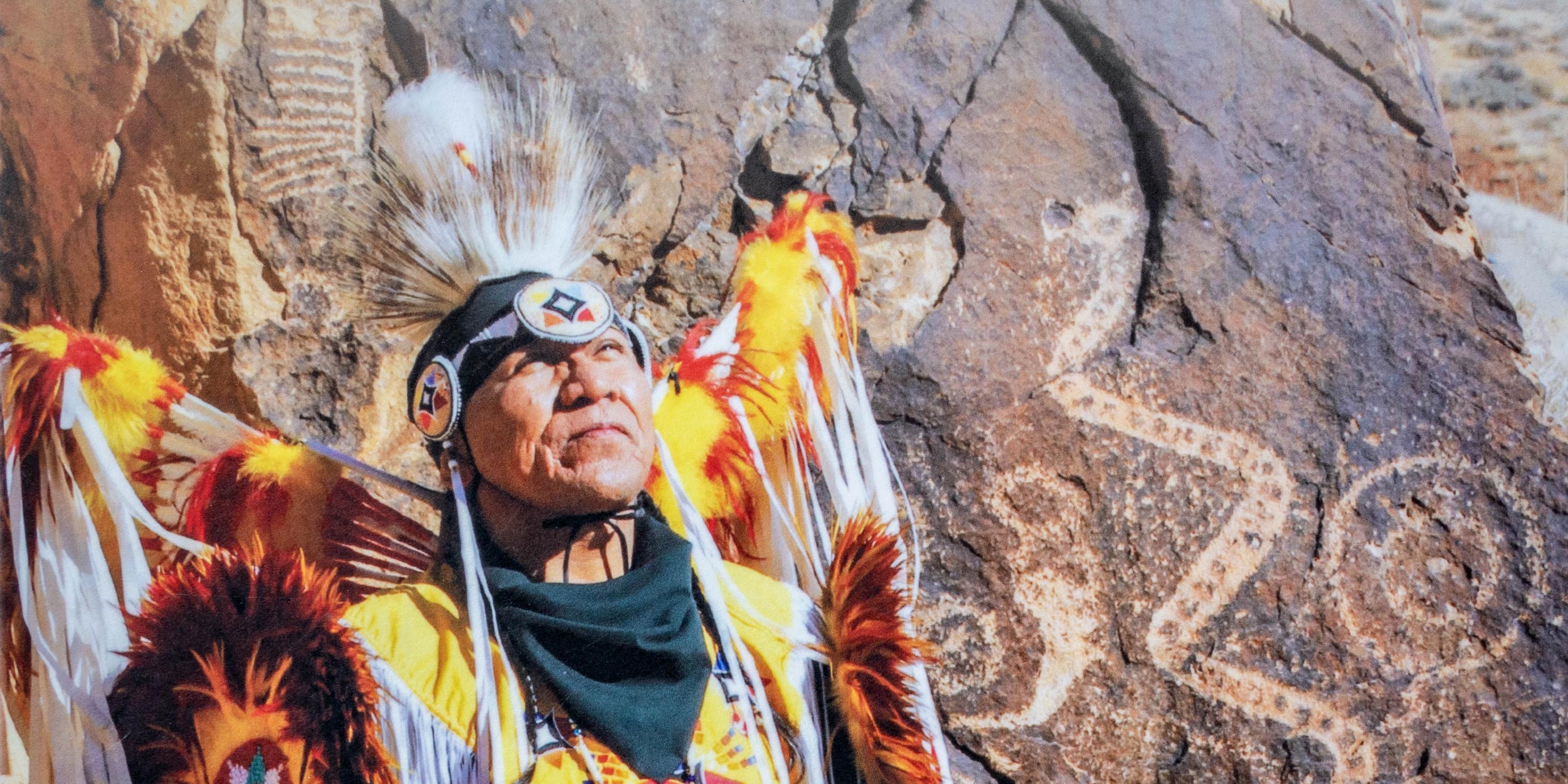 - EXP Journeys A man in traditional Native American attire stands in front of a rock with ancient inscriptions.