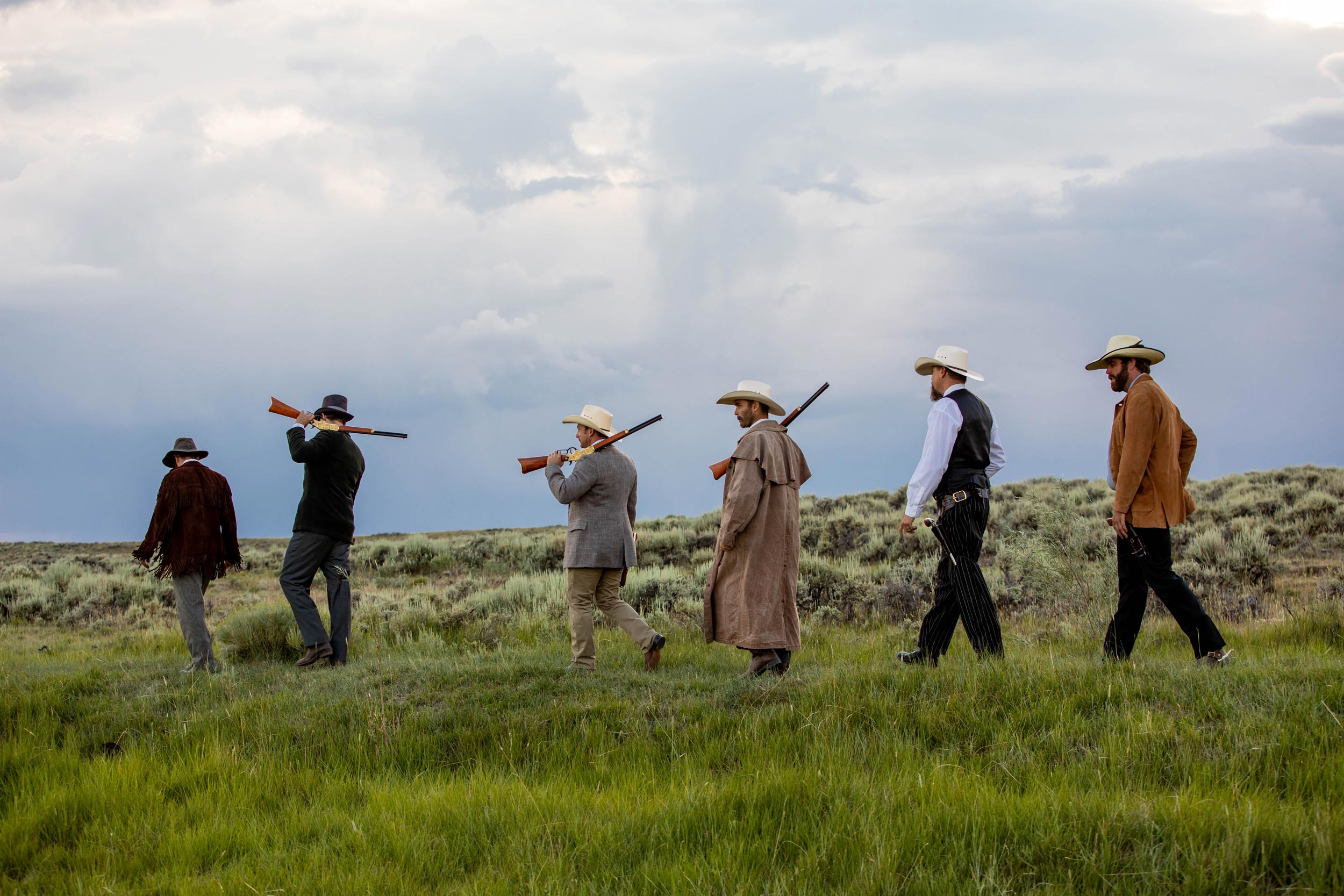 - EXP Journeys Men dressed in wild west clothing walk through grass with guns on their shoulders.