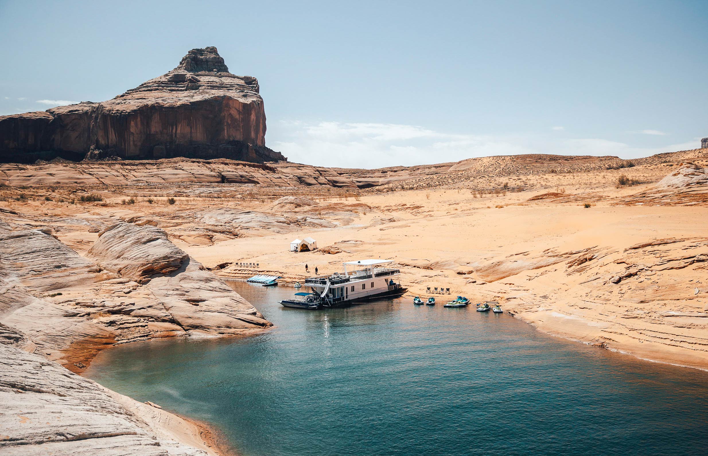 A houseboat sits on the shore of Lake Powell with jet skis and paddle boards ready for guests to enjoy.