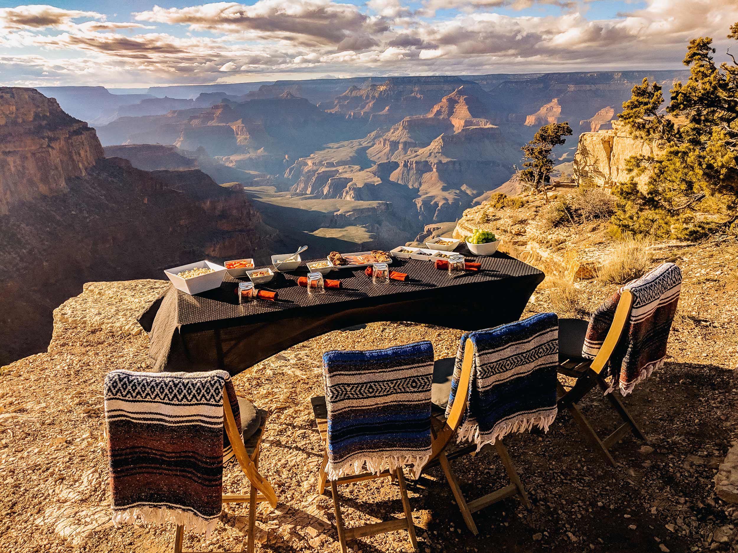 A picnic is set up on a table overlooking the Grand Canyon with sun rays coming through the clouds.