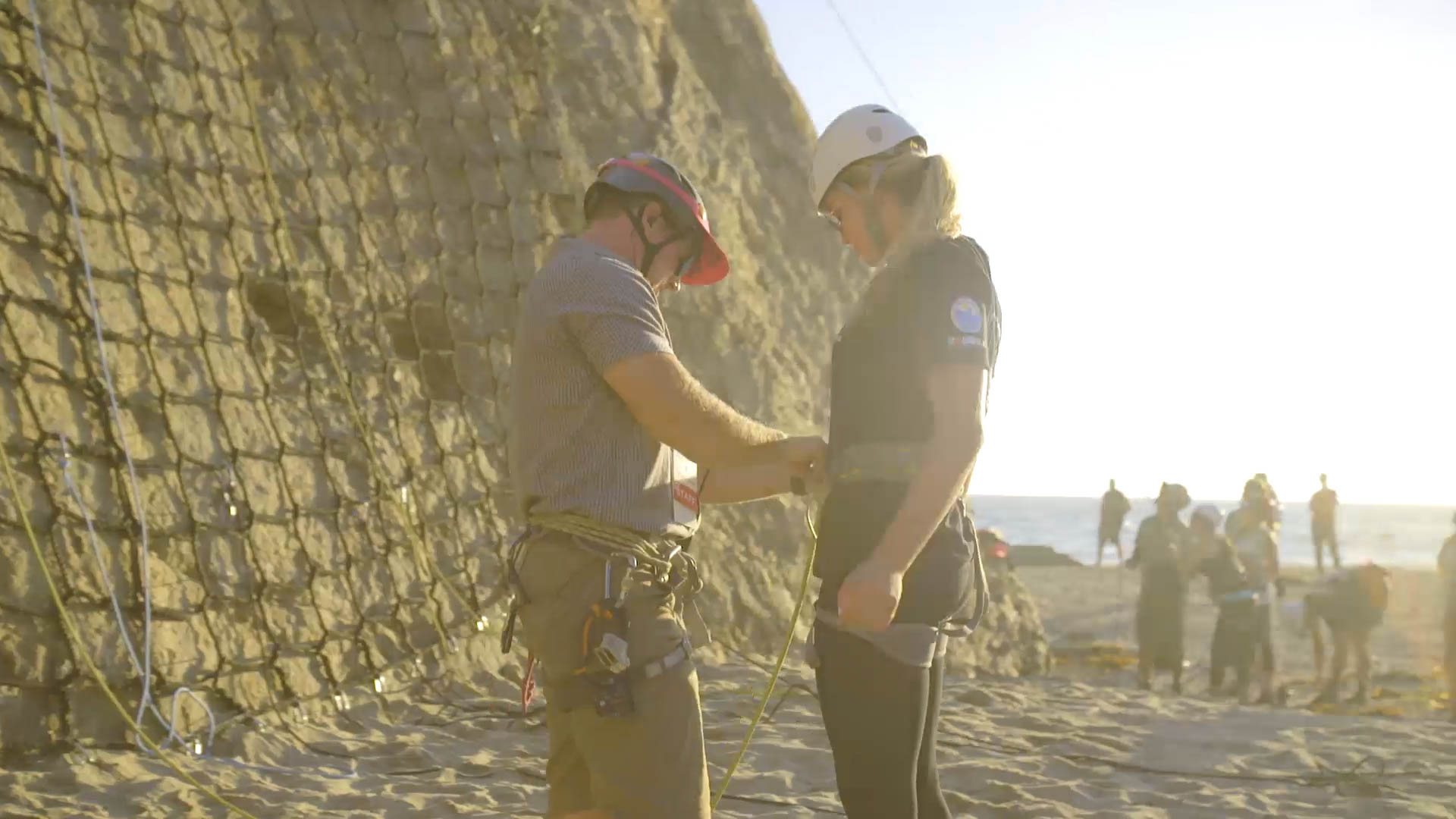 A man puts a harness on a woman to climb up cliffs overlooking the Pacific Ocean.