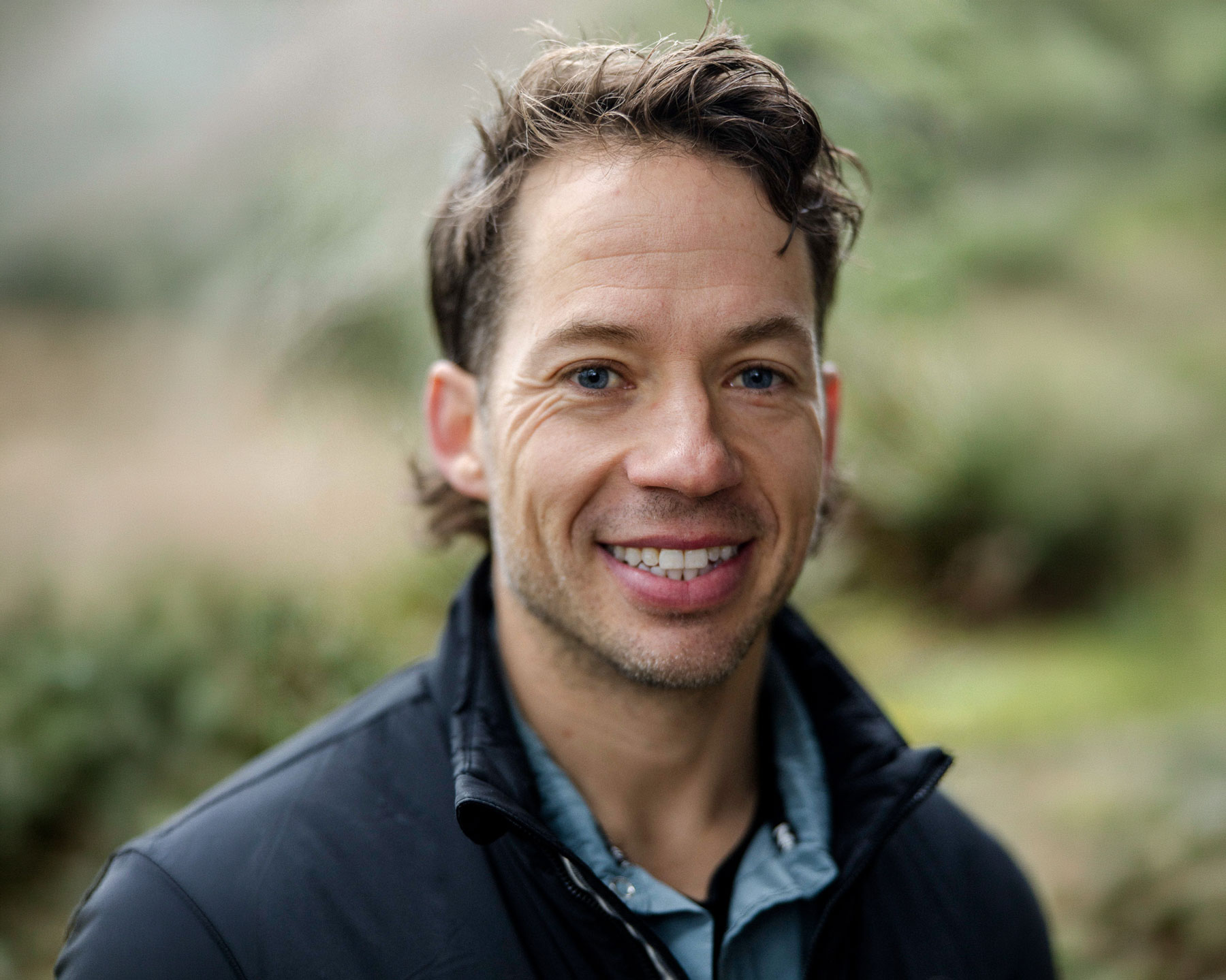 Andrew-Levitt - EXP Journeys Smiling man outdoors, close-up of face with blue eyes and dark jacket.