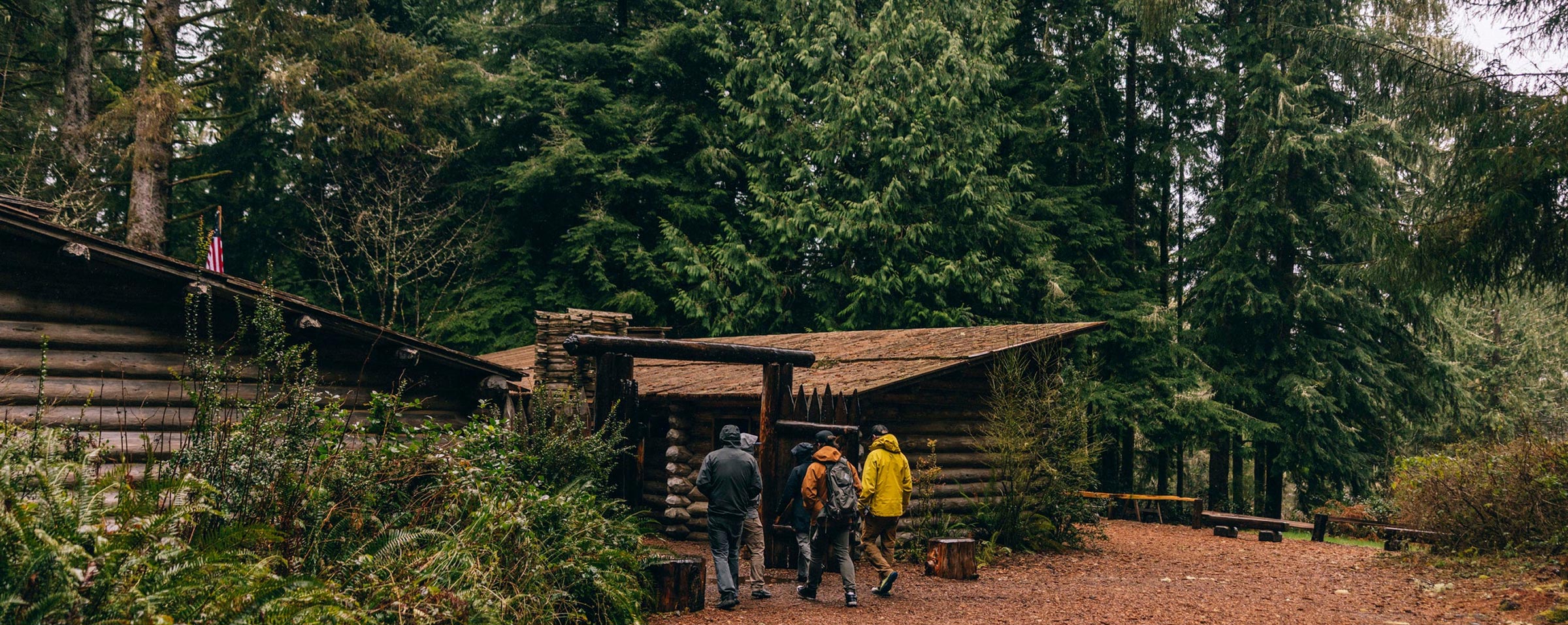 EXP-Guide-School-Hero - EXP Journeys - EXP Journeys Group of hikers in jackets walking toward a rustic log cabin in a dense forest setting.