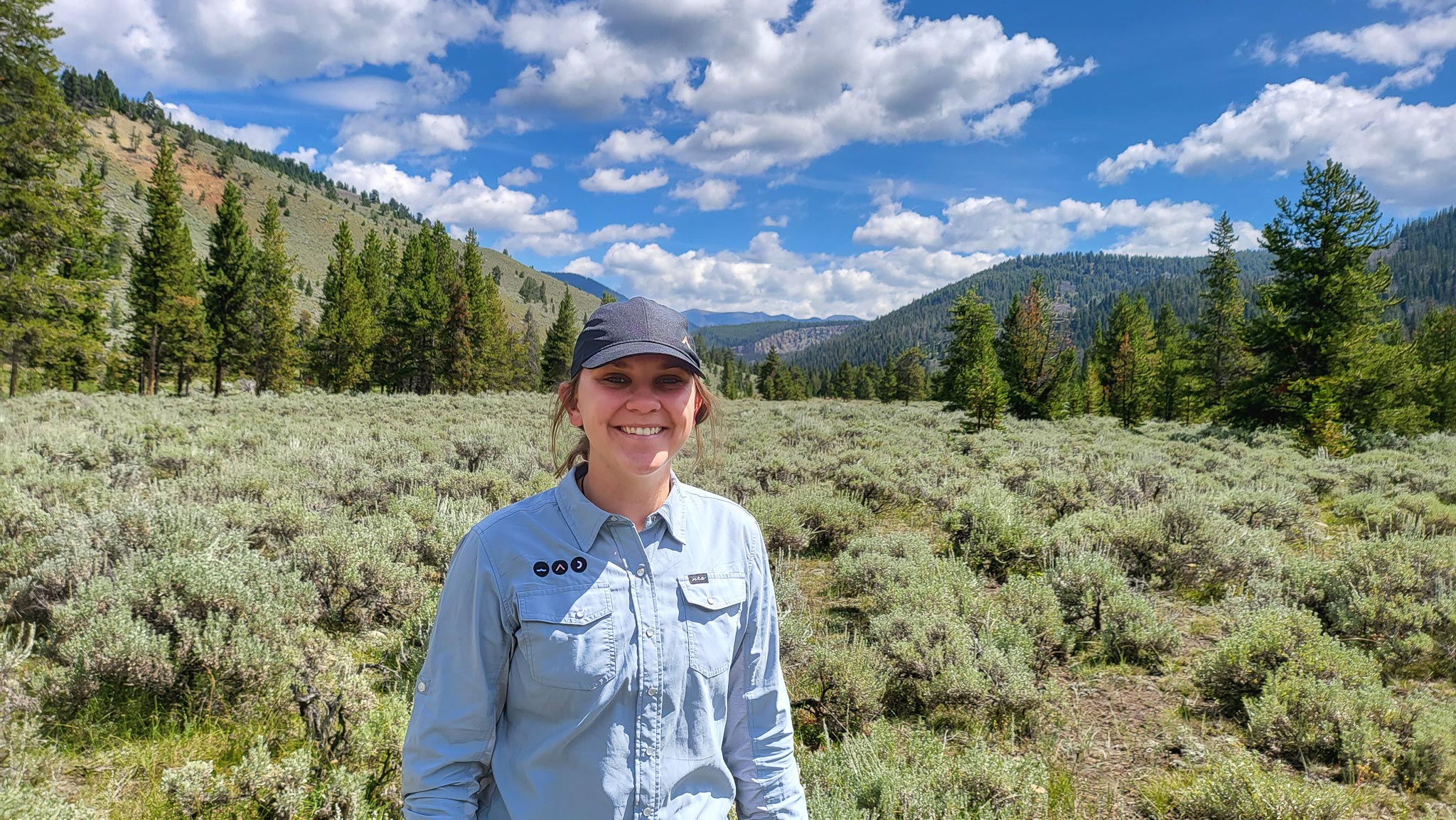 Kathy-Powell - EXP Journeys Smiling woman in a blue button-up shirt and cap stands in a sagebrush field with evergreen trees and mountains in the background under a blue sky.