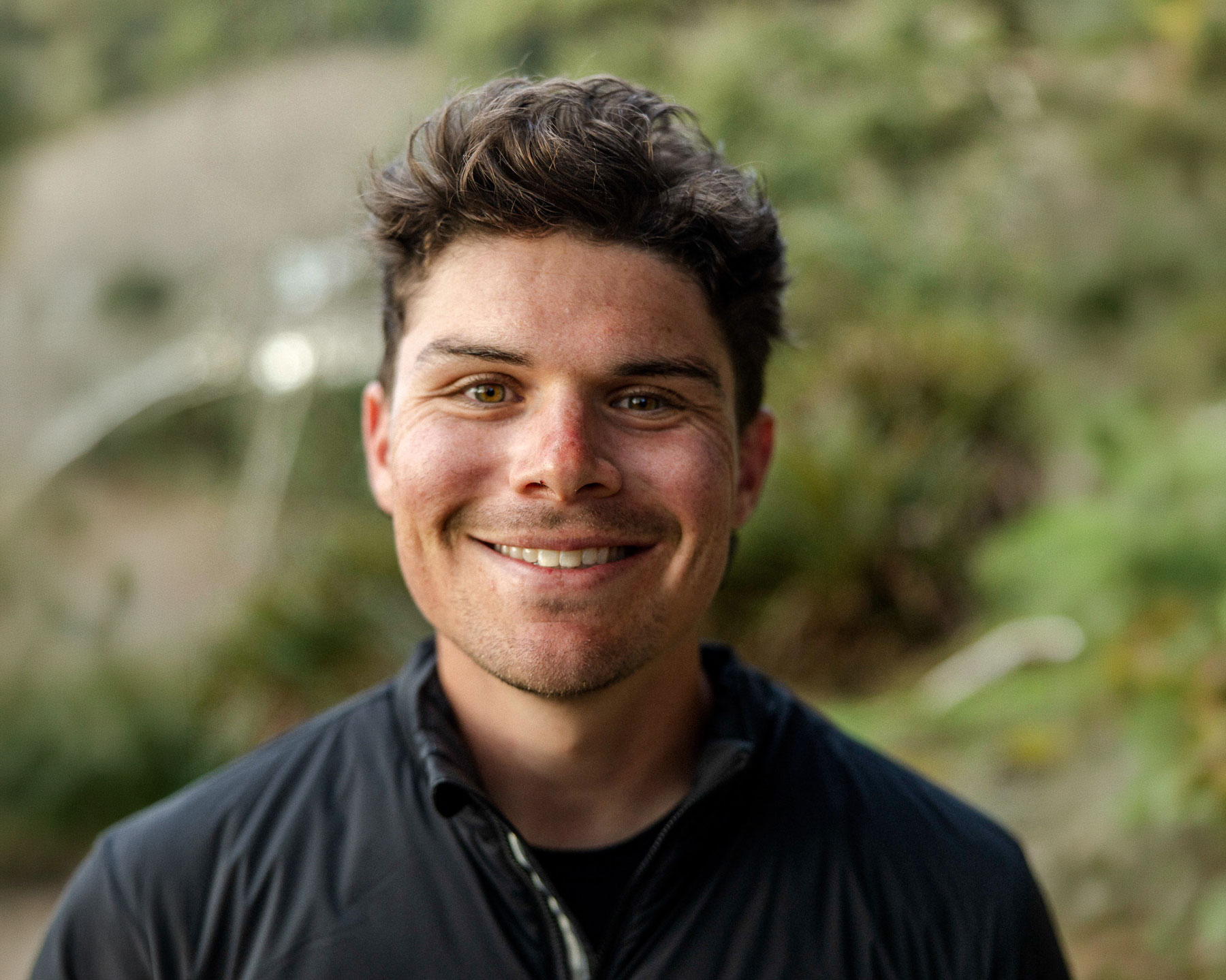 Kyle-Pacioni - EXP Journeys Smiling man with short dark hair outdoors, wearing a dark jacket, facing the camera.