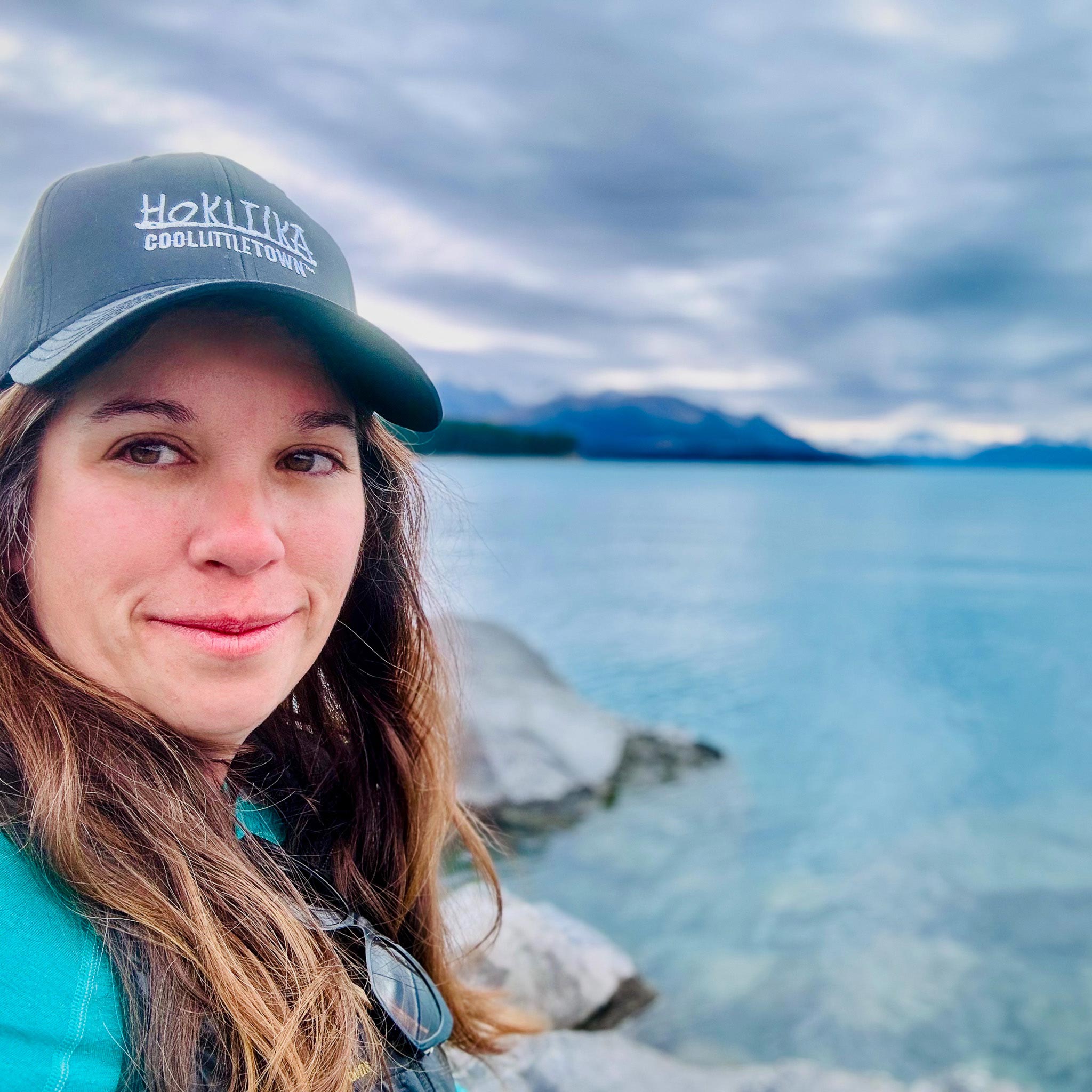 Marie-Hernandez - EXP Journeys Partial portrait of a woman wearing a teal jacket and gray cap by a lake with mountains in the background.