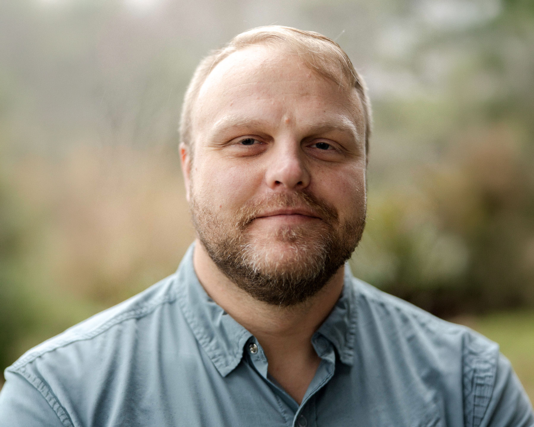 Mitch-McElwain - EXP Journeys Close-up portrait of a bearded man in a light blue shirt outdoors, smiling softly with a blurred natural background.