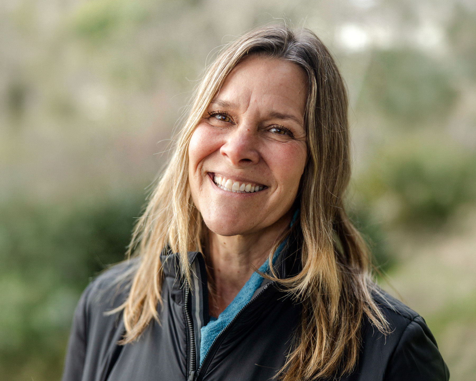 Rene-Collins - EXP Journeys Smiling woman with long blond hair wearing a black jacket, outdoors with a blurred green background