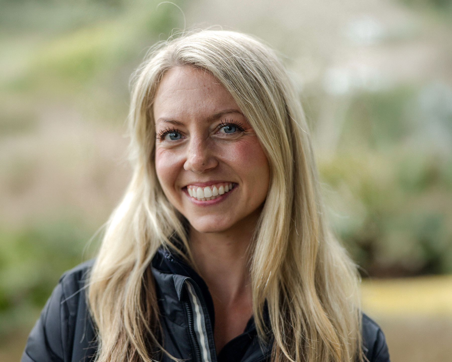 Sarah-Webber - EXP Journeys Close-up portrait of a smiling blonde woman outdoors with a blurred natural background.