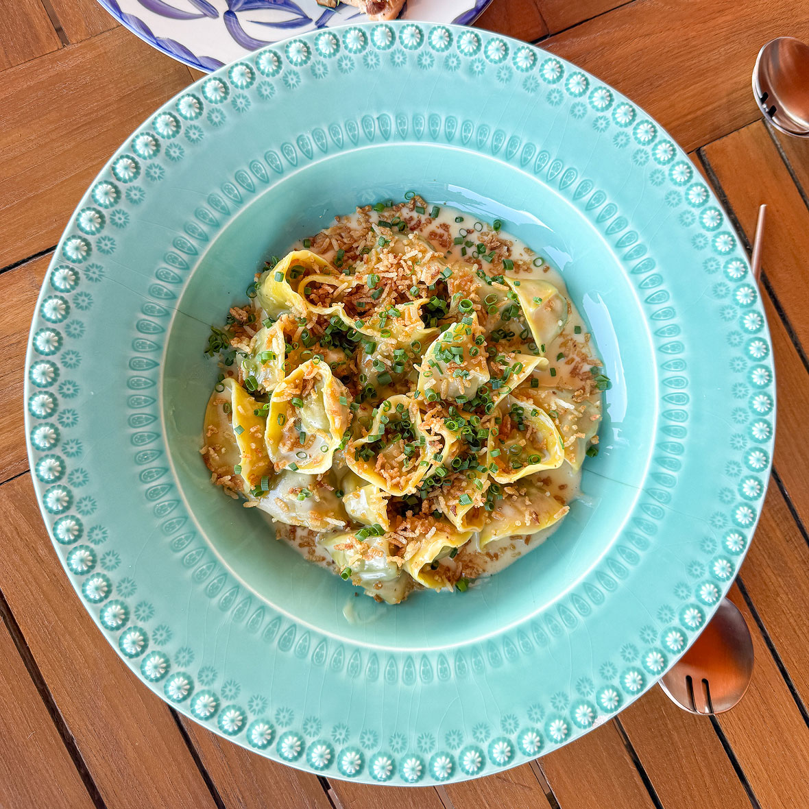 Teal decorative plate with creamy tortellini, crispy onions, and chopped chives on a wooden table.