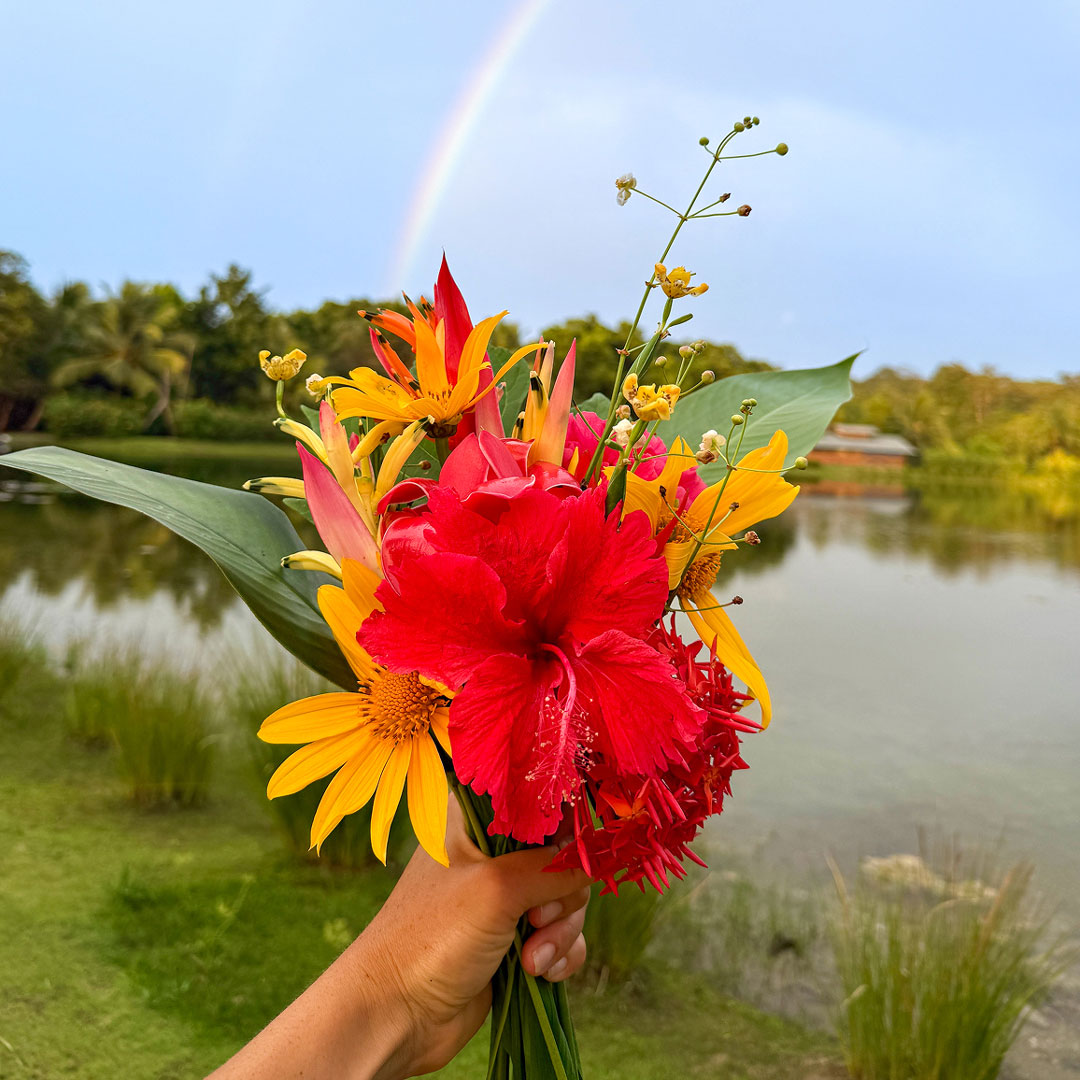 Bright bouquet of red, yellow, and orange flowers held in a hand, with a rainbow arcing over a calm lake in the background.