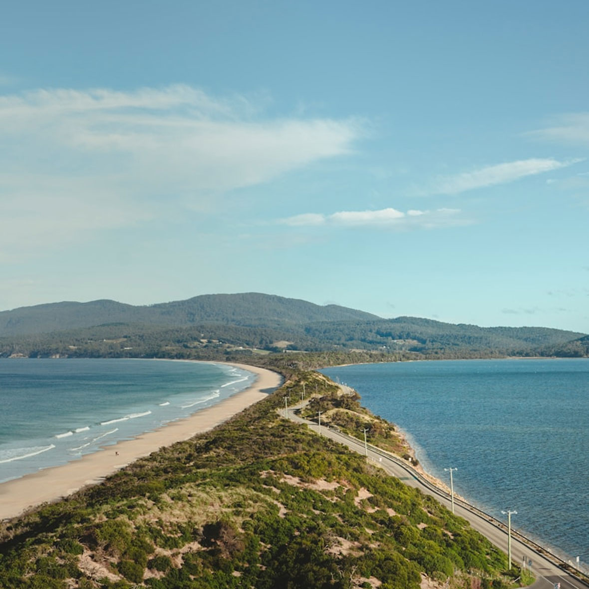 Australia-Bruny-Island - EXP Journeys - EXP Journeys Narrow coastal spit with a road running along its edge, flanked by blue ocean on one side and a sandy beach on the other, hills in the distance under a clear sky.