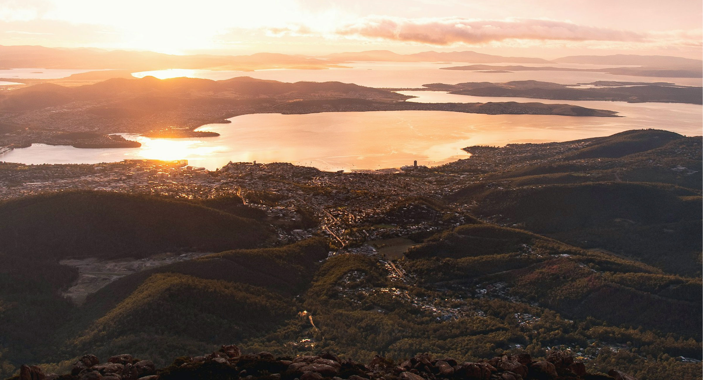 Australia-Hobart - EXP Journeys - EXP Journeys Aerial view of a coastal city at sunset, with golden water and surrounding islands in the distance to the horizon.