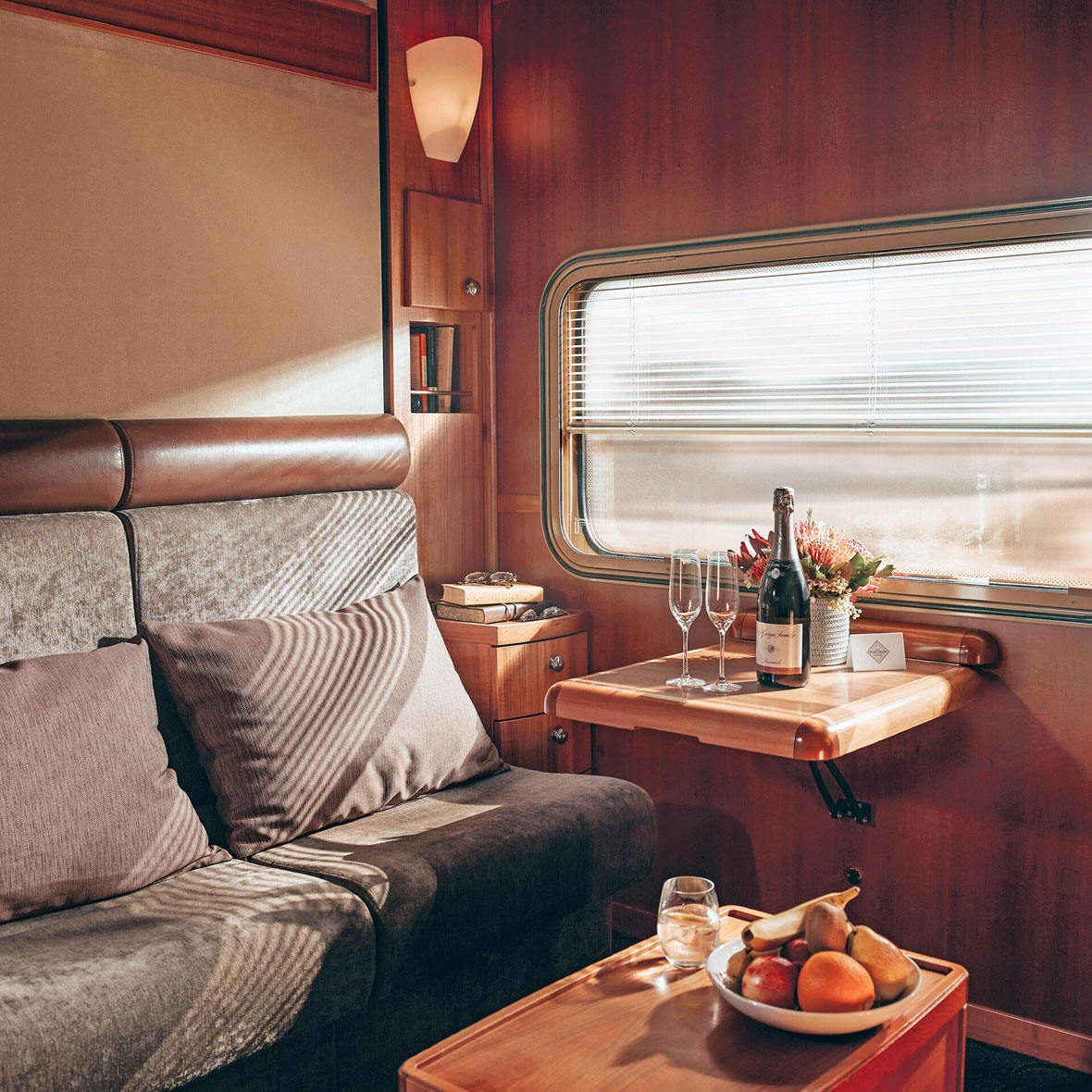 Australia-Indian-Pacific-Train-Interior - EXP Journeys - EXP Journeys Cozy train cabin with a gray sofa, cushions, and a wood-paneled wall by a window with blinds.