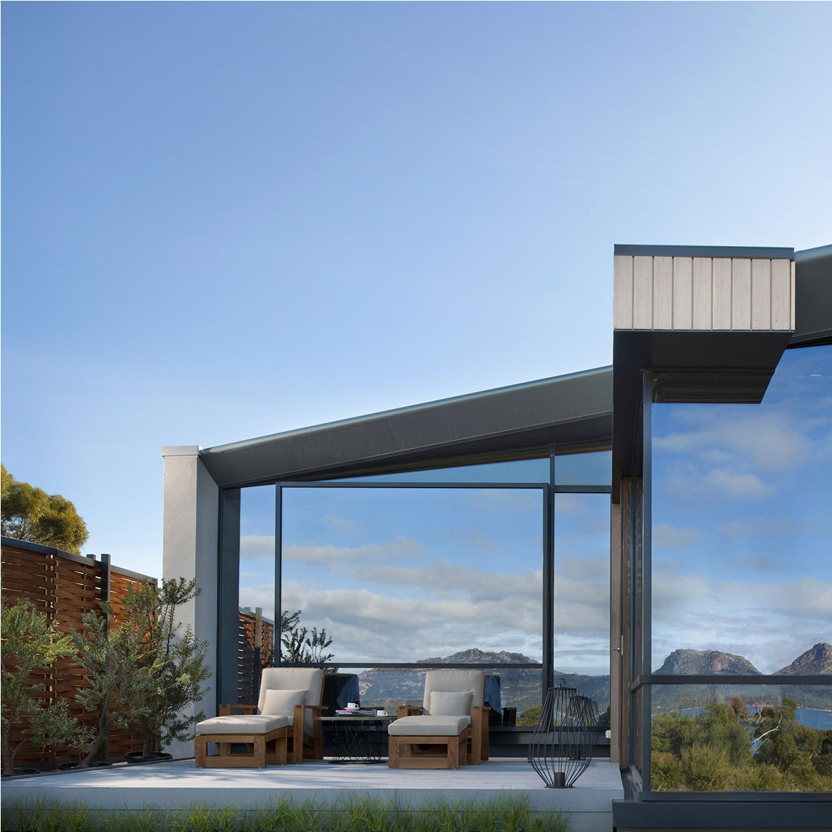 Australia-Saffire-Freycinet-Exterior - EXP Journeys - EXP Journeys Modern glass-walled patio with wooden lounge chairs on a light deck, overlooking distant mountains under a clear blue sky.