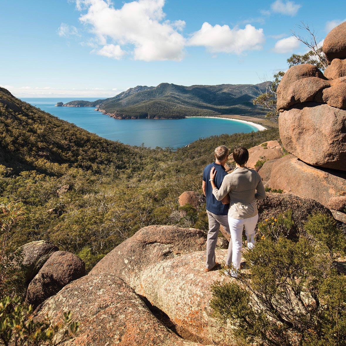 Australia-Saffire-Freycinet-Wineglass-Bay - EXP Journeys - EXP Journeys Couple standing on a rocky overlook, watching a crescent beach and blue bay surrounded by hills.