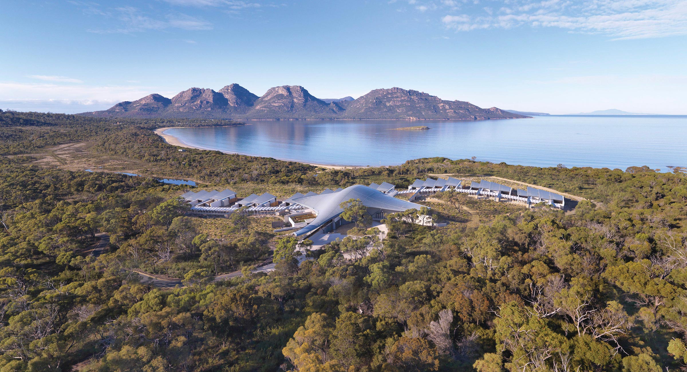 Australia-Saffire-Freycinet - EXP Journeys - EXP Journeys Aerial view of a coastal landscape with a curved-roof modern resort surrounded by trees and a calm blue bay.
