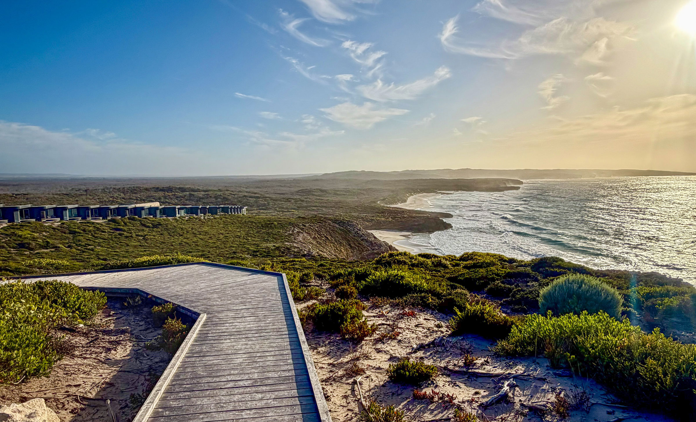 Australia-Southern-Ocean-Lodge - EXP Journeys - EXP Journeys Wooden boardwalk stretching toward a sandy beach, with low coastal shrubs and the ocean to the right under a clear blue sky, sun bright overhead.