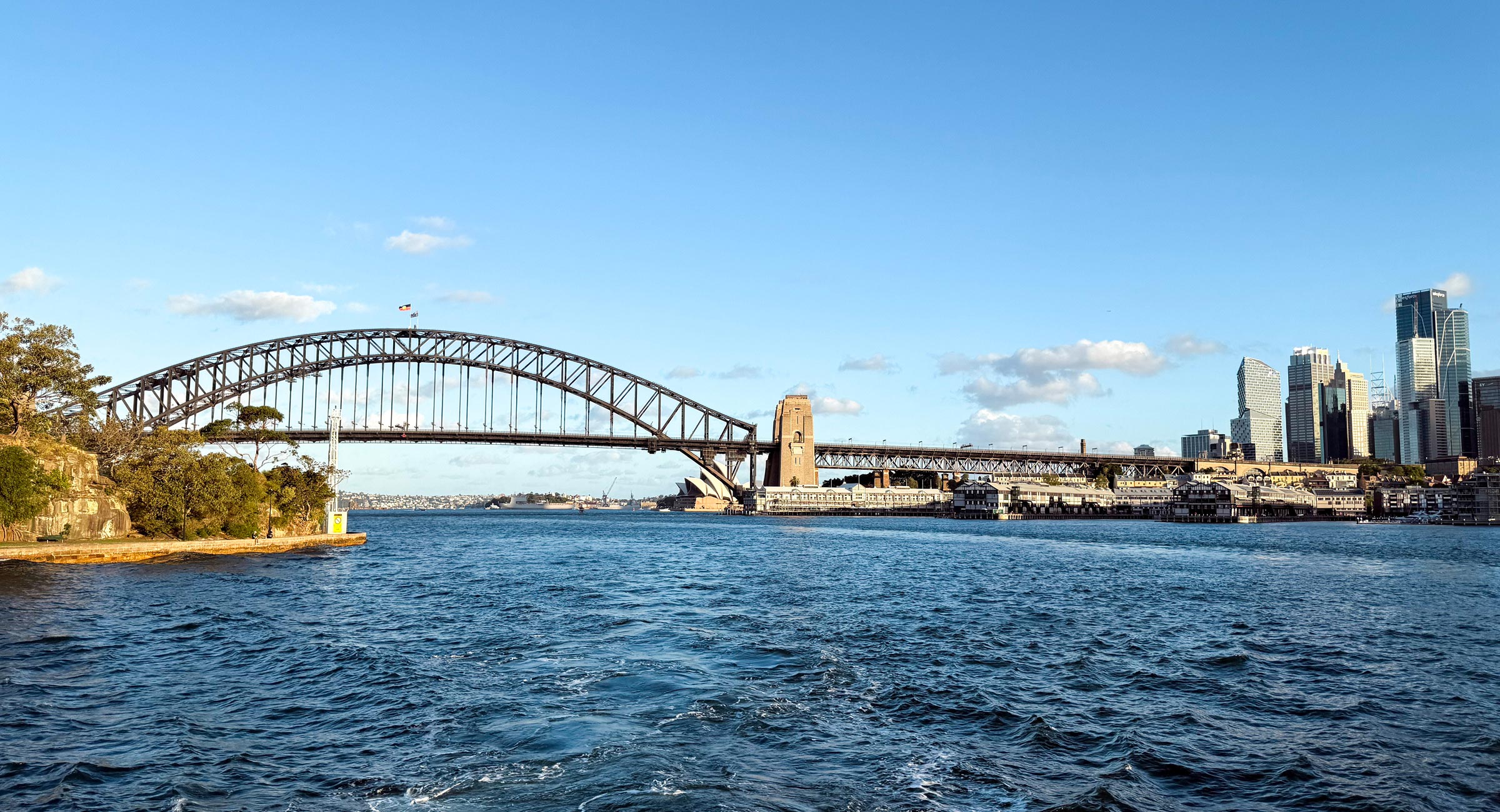 Australia-Sydney-Sunset-Cruise - EXP Journeys - EXP Journeys Sydney Harbour Bridge and skyline viewed from the harbor on a clear day, with blue water in the foreground.