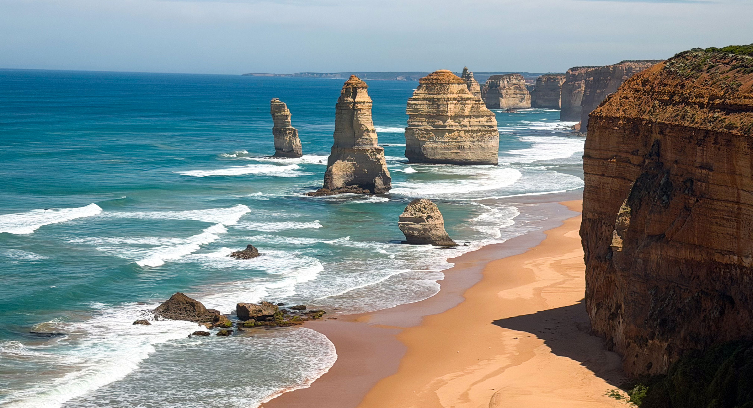 Australia-The-Twelve-Apostles - EXP Journeys - EXP Journeys Coastal scene with limestone sea stacks rising from turquoise waves along a sandy beach and cliffs under a clear sky.