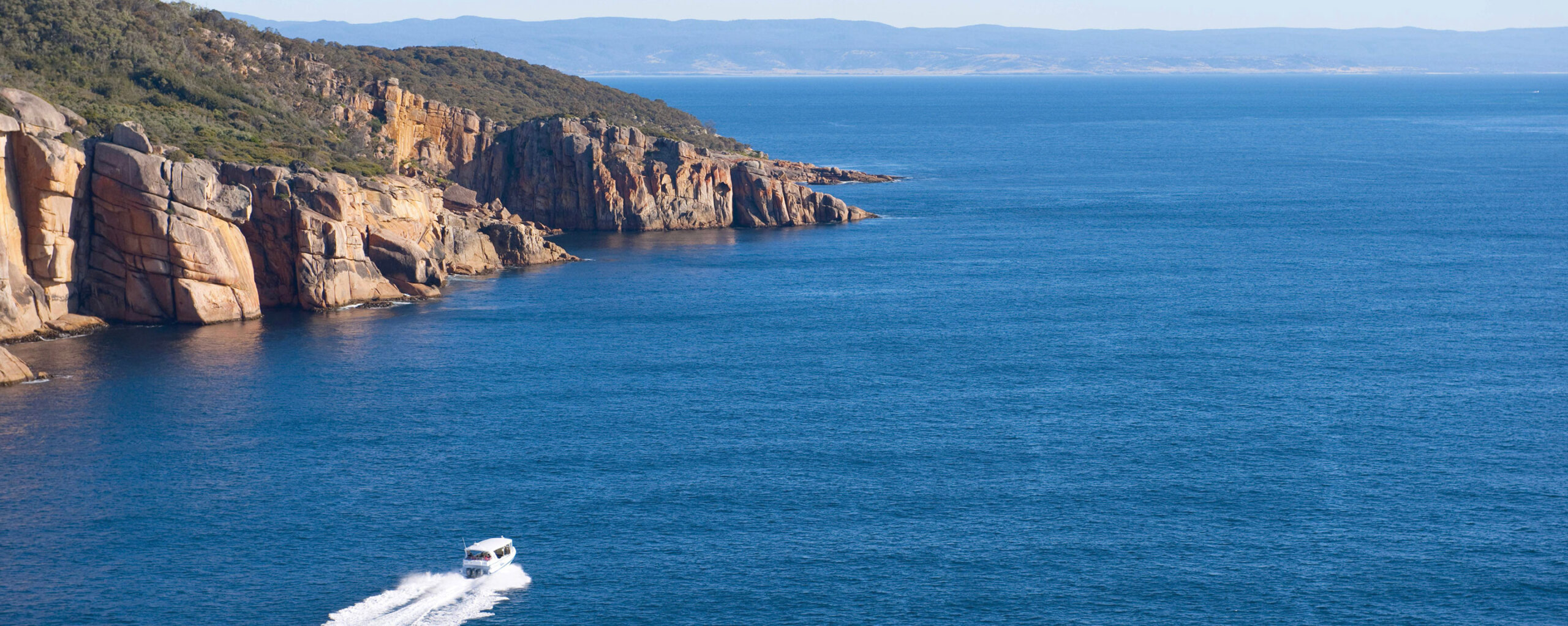 EXP-Journeys-Deeper-Down-Under-A-Family-Adventure-Through-Australia - EXP Journeys - EXP Journeys Rugged rocky coastline on the left with calm blue water and a small speedboat in the foreground left creating a wake.