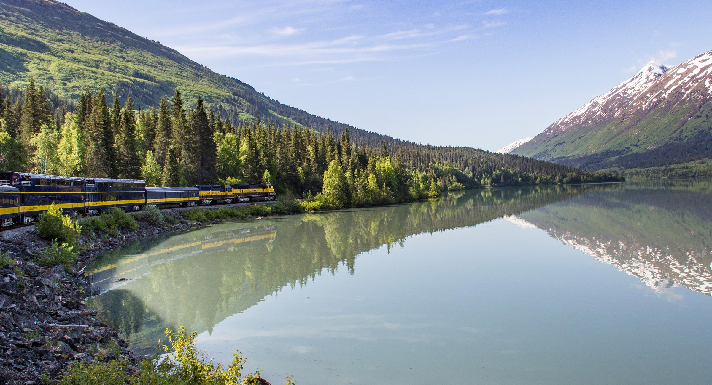 Alaska-Denali-Star-Train - EXP Journeys - EXP Journeys Train on a blue-and-yellow locomotive line travels along a calm lake with evergreen trees and snow-capped mountains in the background far to the right.