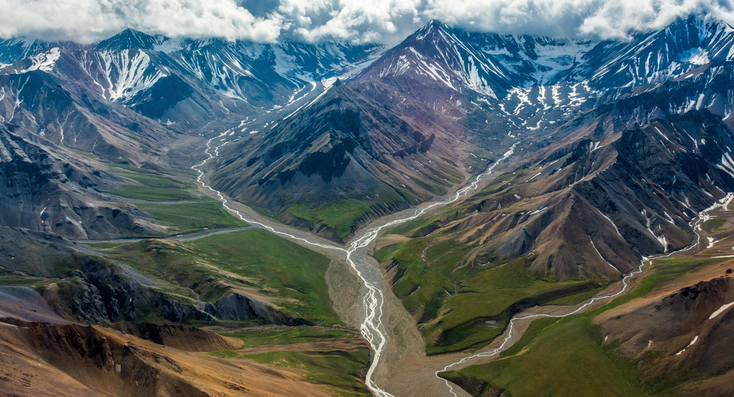 Alaska-Denalil - EXP Journeys - EXP Journeys Aerial view of a rugged mountain range with snow patches and braided streams winding through green valleys below.