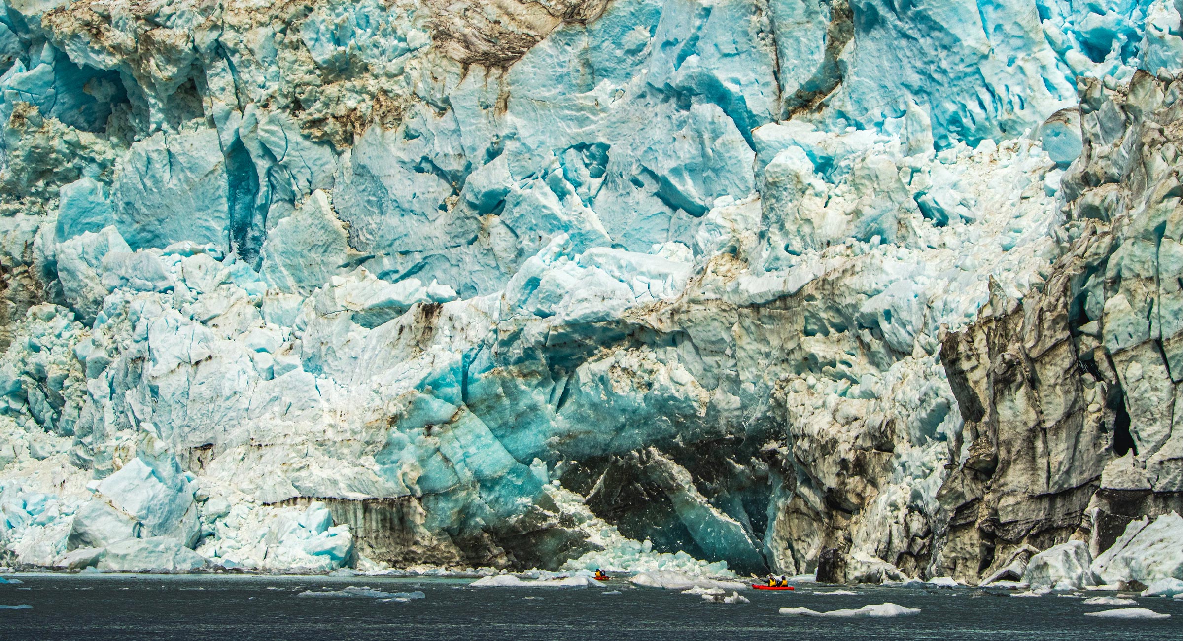 Alaska-Glacier-Bay-private-kayak-tour - EXP Journeys - EXP Journeys Massive blue ice glacier wall with jagged surfaces towering over dark water; two small red boats with people at the shoreline for scale