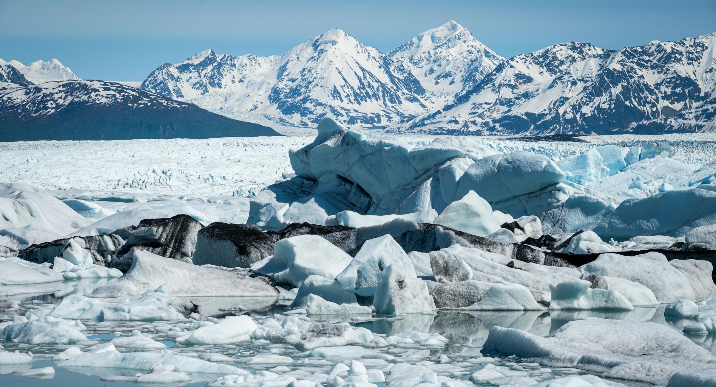 Alaska-Knik-Glacier - EXP Journeys - EXP Journeys Wide view of a frozen glacier with blue-tinged ice chunks and snow-covered mountains under a clear blue sky, reflecting in calm water.