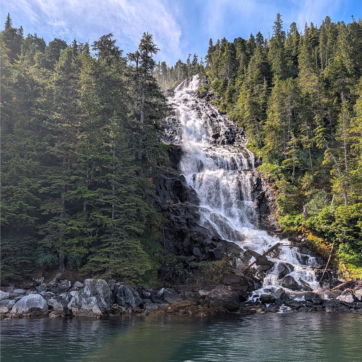 Alaska-luxury-yacht-tour-waterfall - EXP Journeys - EXP Journeys Waterfall cascading down a rocky cliff into a calm lake surrounded by evergreen trees in a forest setting.