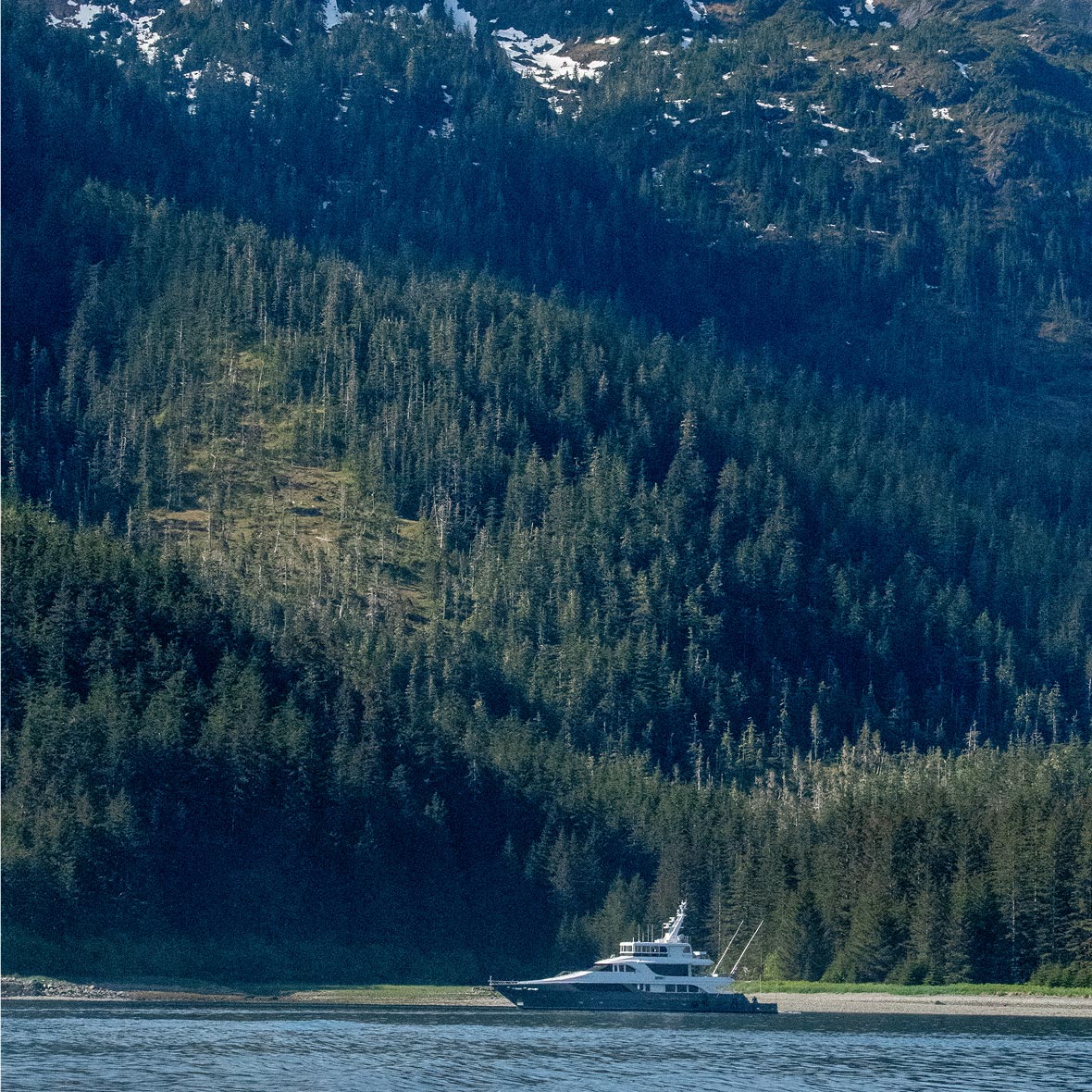 Alaska-luxury-yacht-tour - EXP Journeys - EXP Journeys A white yacht cruising near a forested shore with tall evergreen trees and snow-capped mountains in the distance.