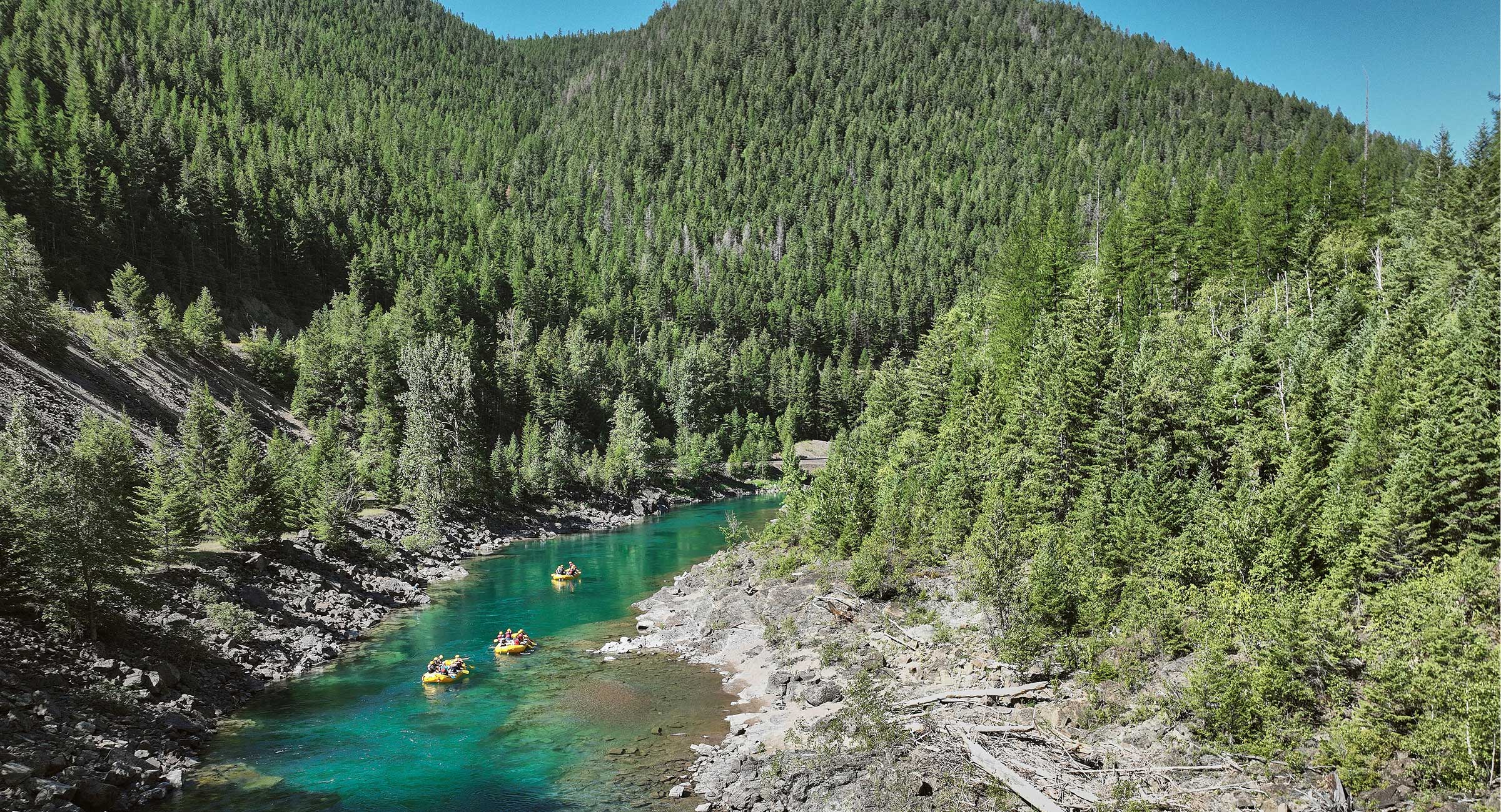 Alaska-whitewater-rafting - EXP Journeys - EXP Journeys Turquoise river winding through a dense evergreen forest; several yellow rafts with paddlers.