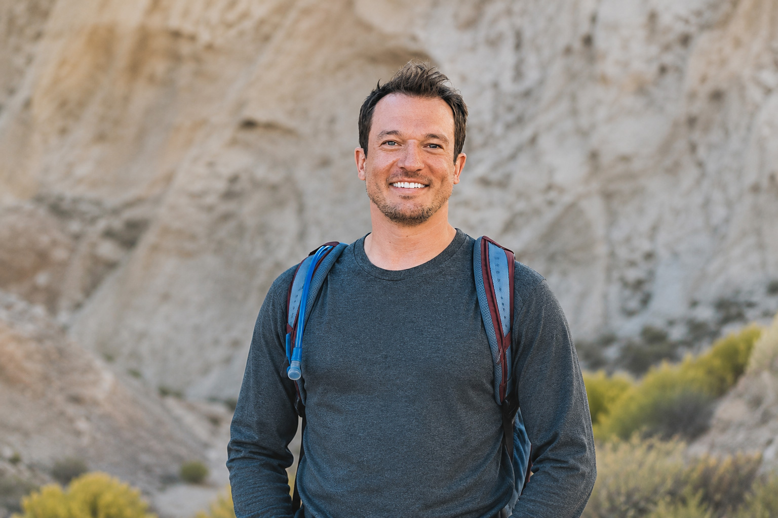 Alex-Yakshe - EXP Journeys Smiling man with a blue and red backpack standing in a rocky desert landscape.